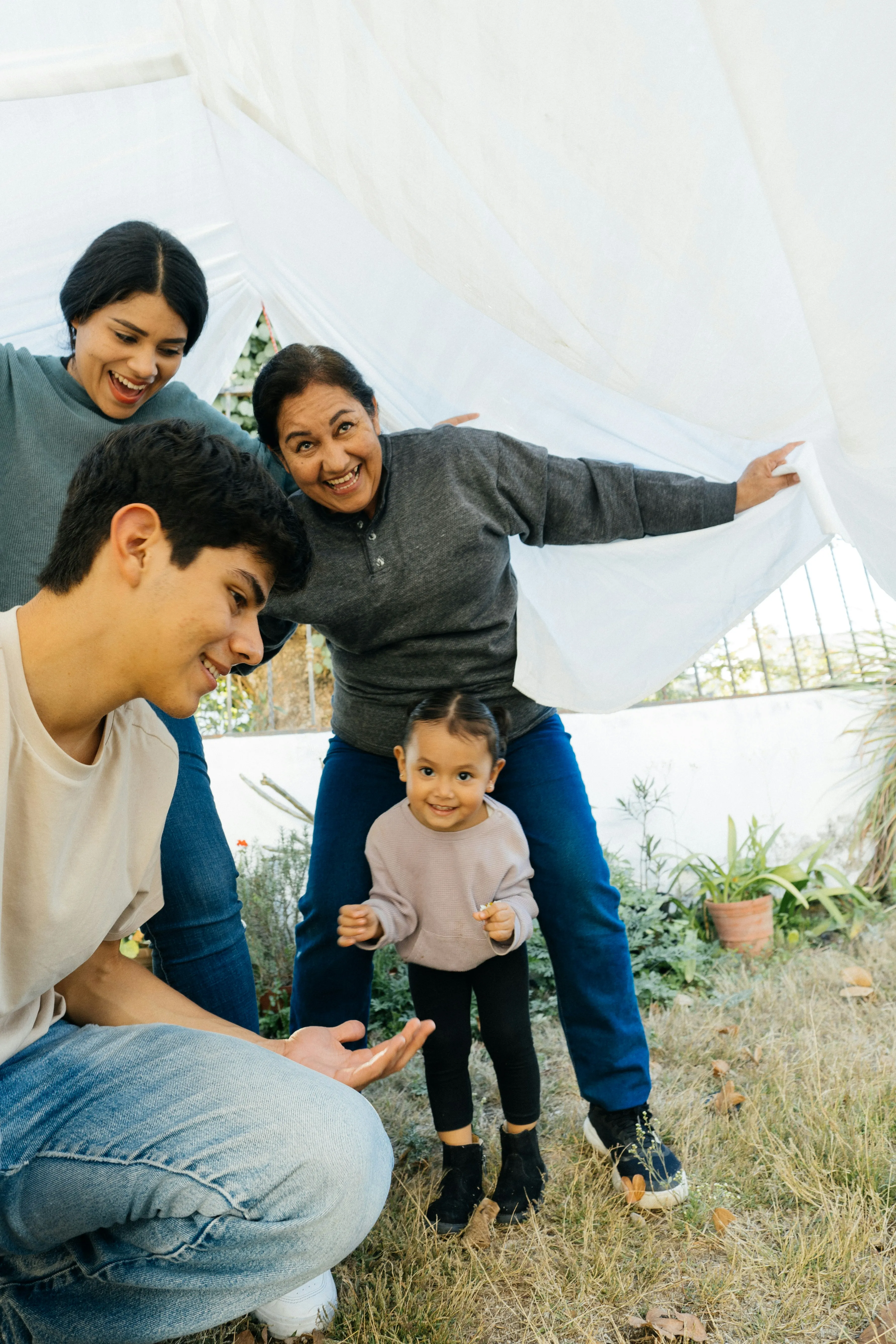 Smiling multigenerational family playing outside under a white sheet in a garden.