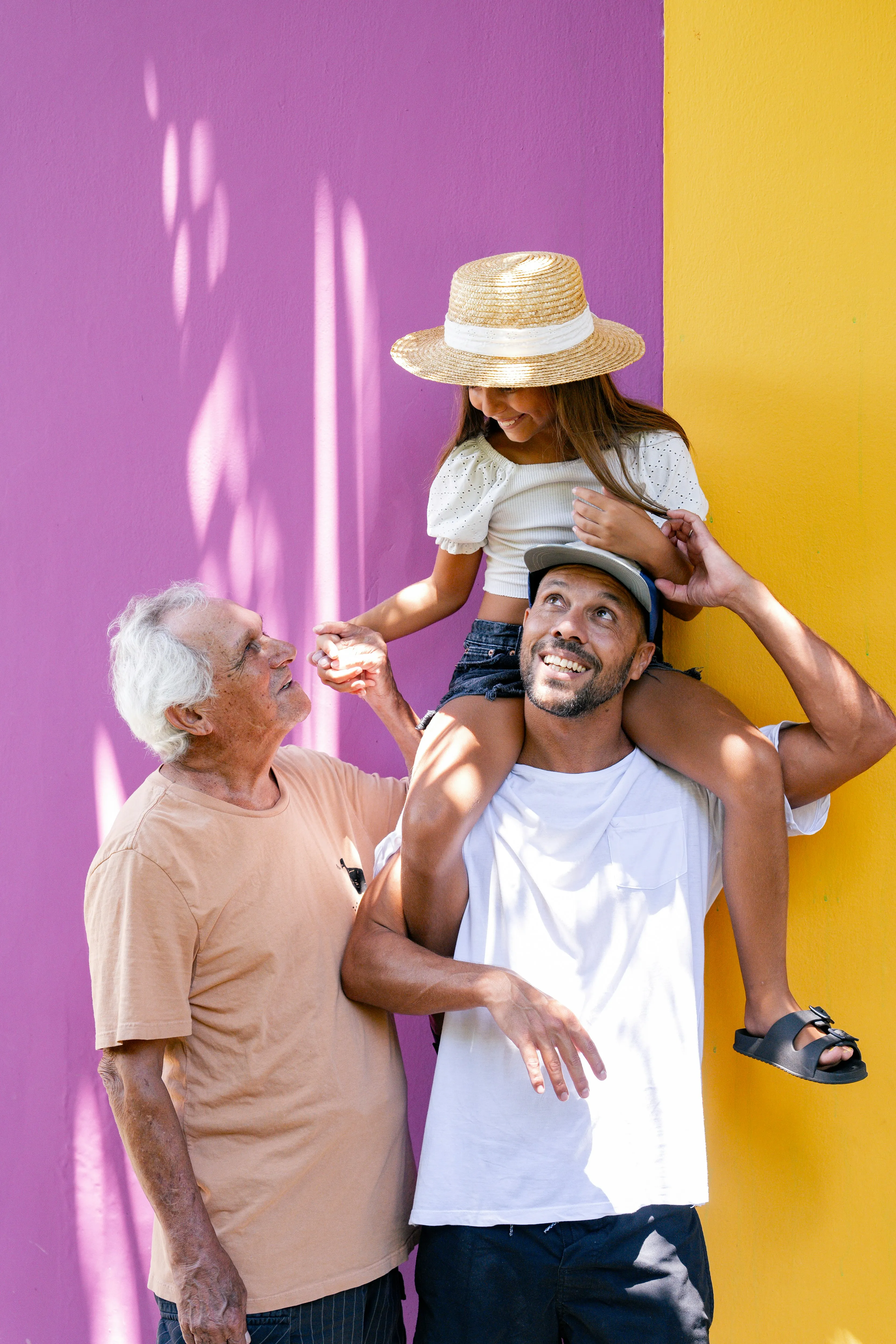 Smiling elderly man holding hands with a man carrying a young girl on his shoulders against a pink and yellow wall.