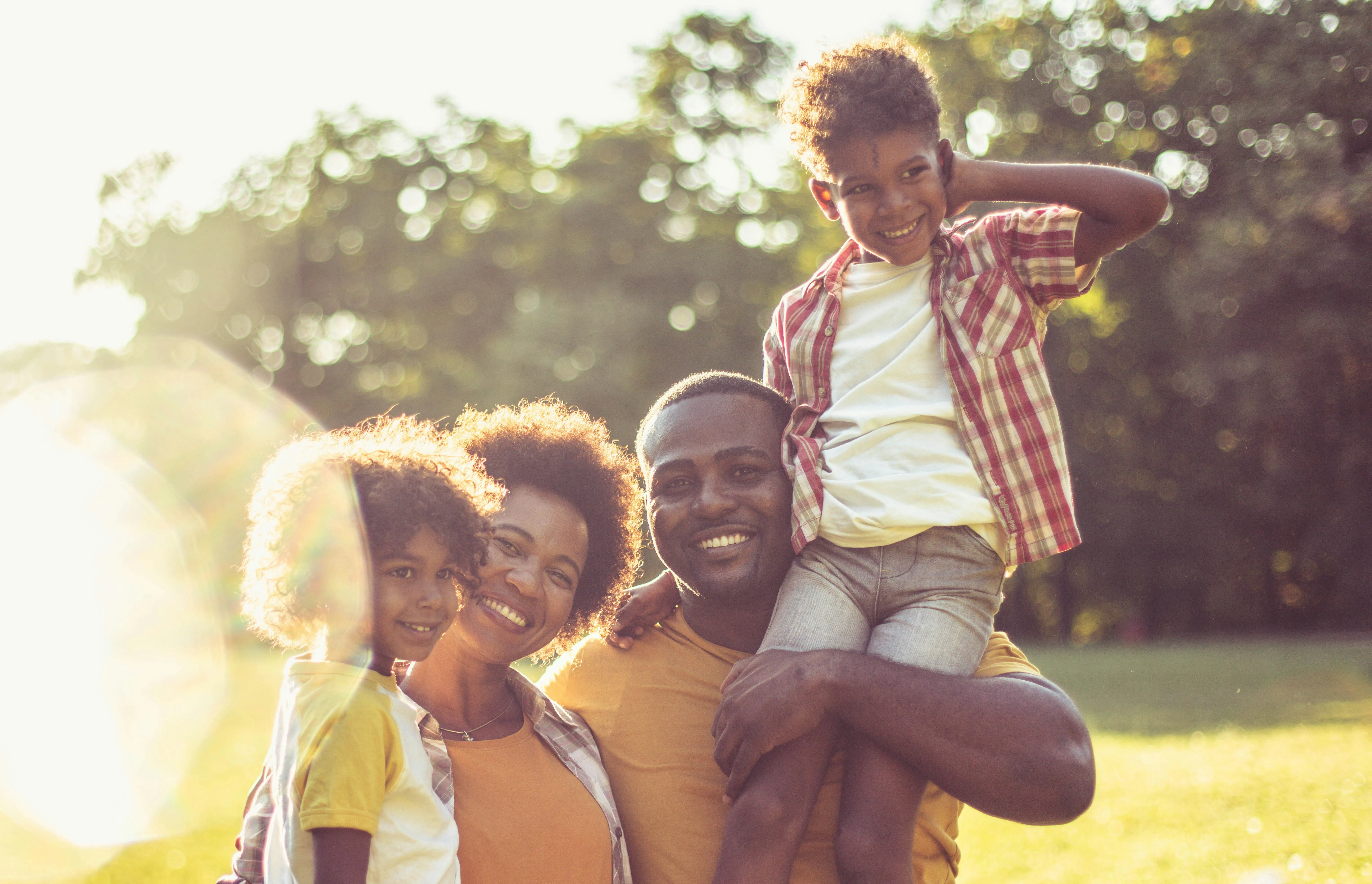 Happy family outdoors with two children; one child sitting on the father's shoulders, all smiling.