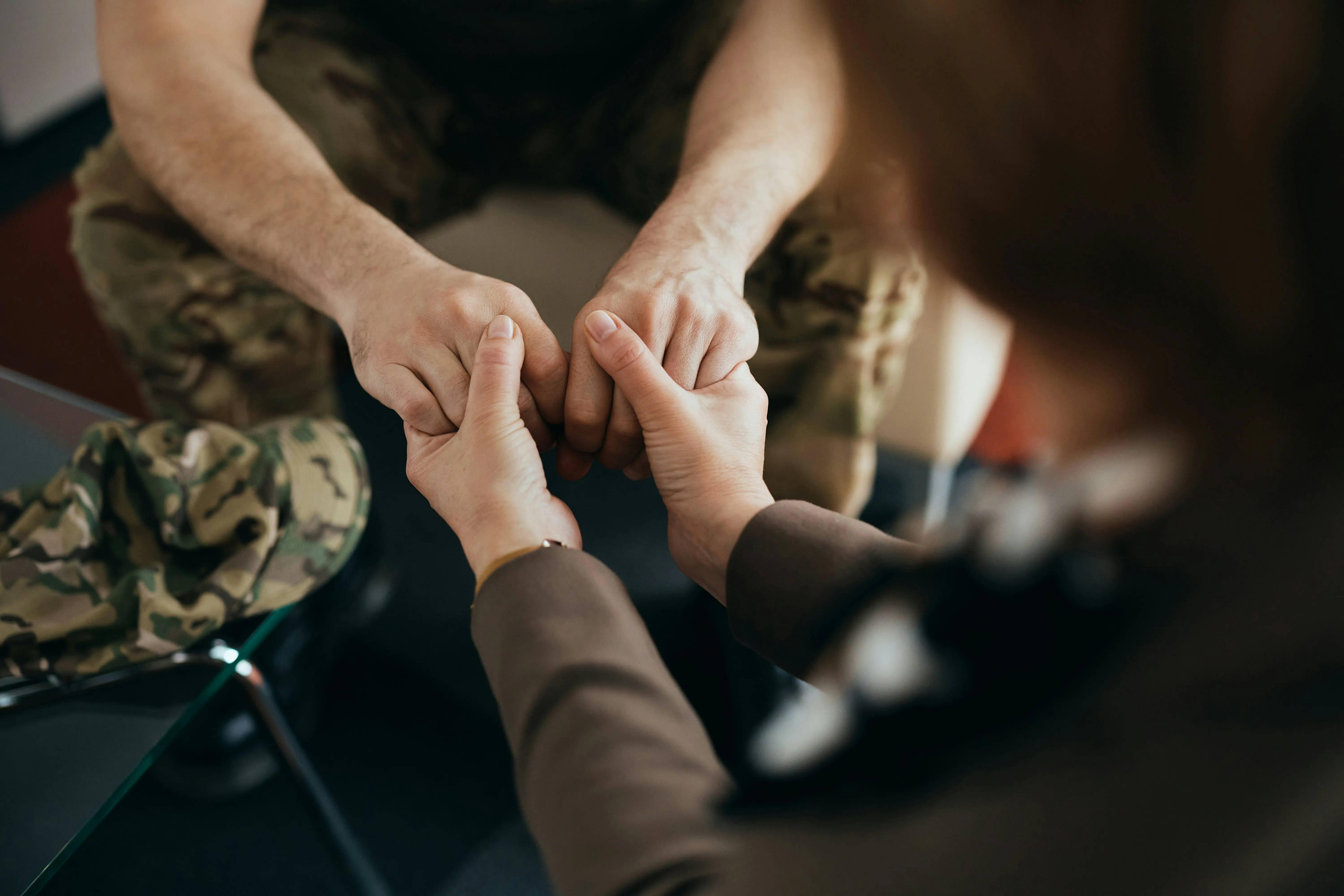 Person in military camouflage holding hands with another person wearing a brown jacket in a supportive gesture.