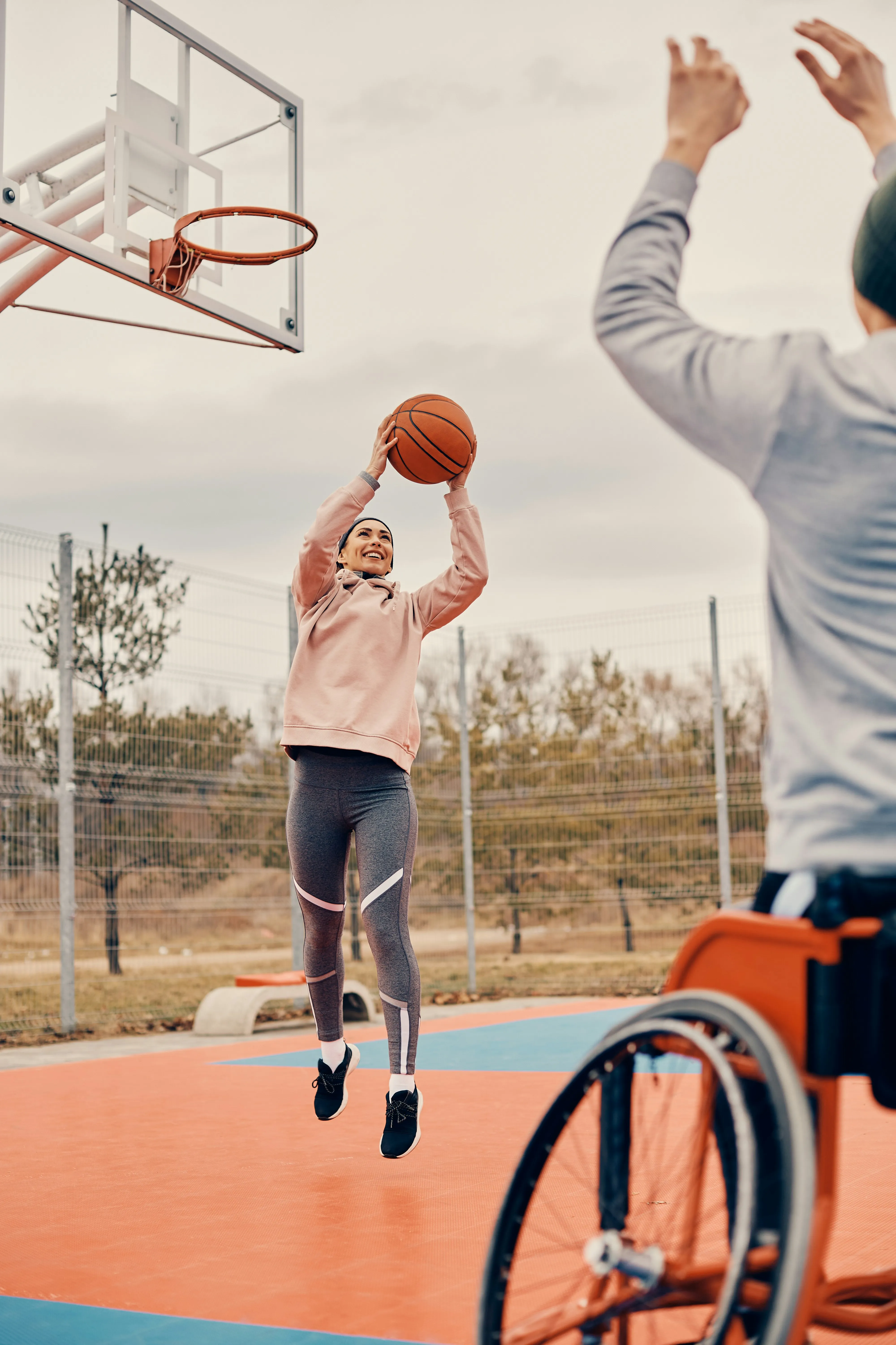 Woman in activewear jumping to shoot a basketball on an outdoor court while a person in a wheelchair raises their hands in defense.