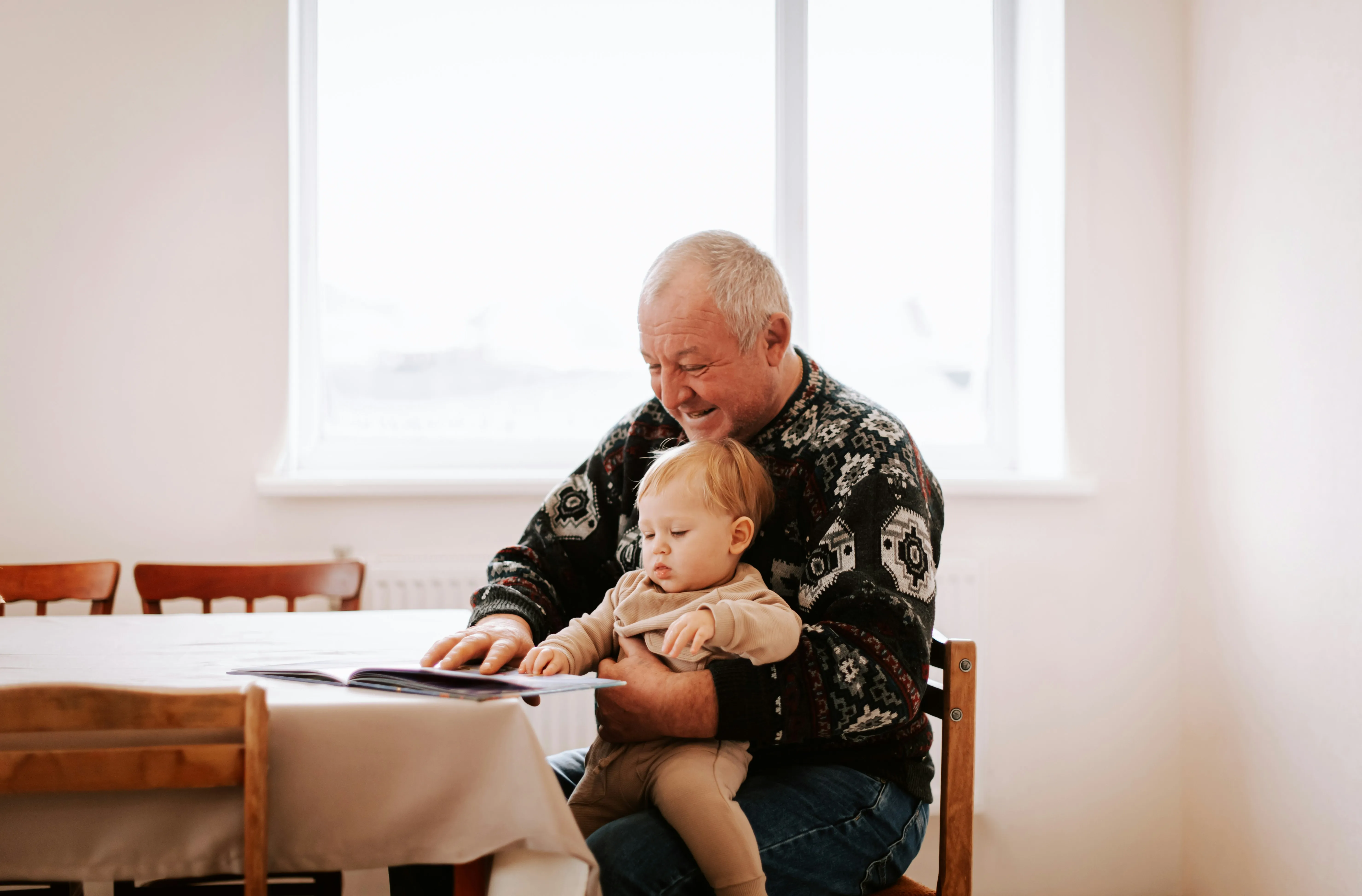 Elderly man reading a book with a toddler on his lap at a dining table in a bright room.