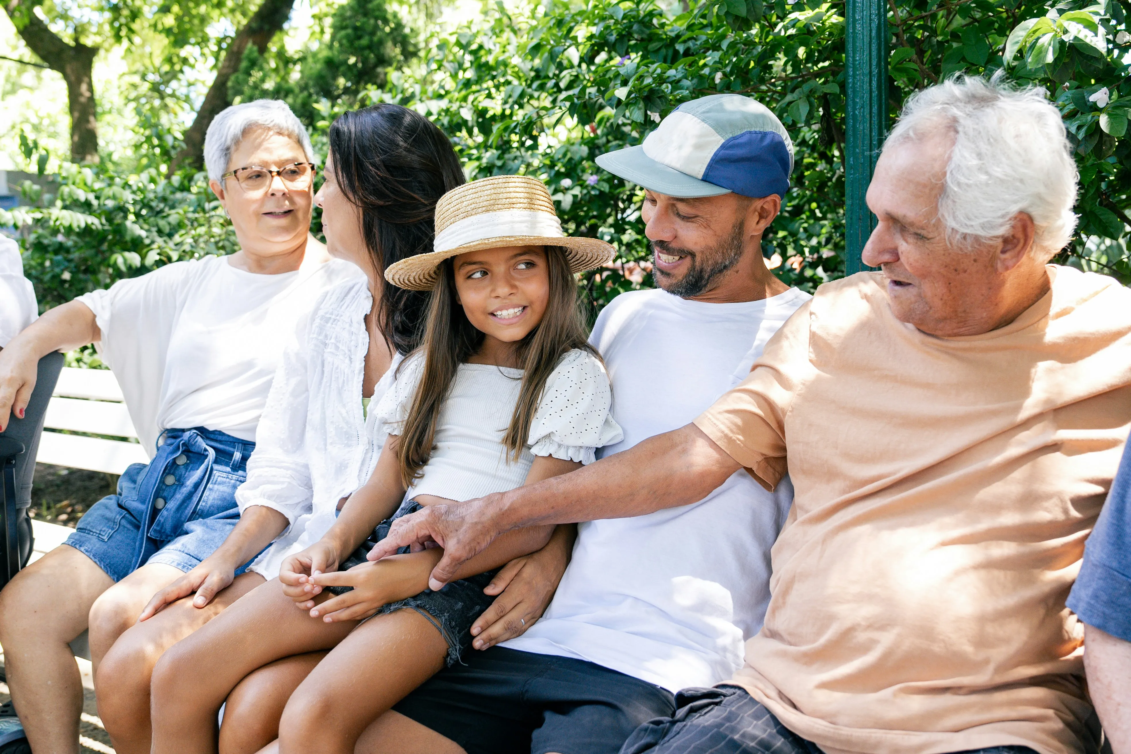 Multigenerational family sitting closely on a bench outdoors, smiling and interacting with each other.