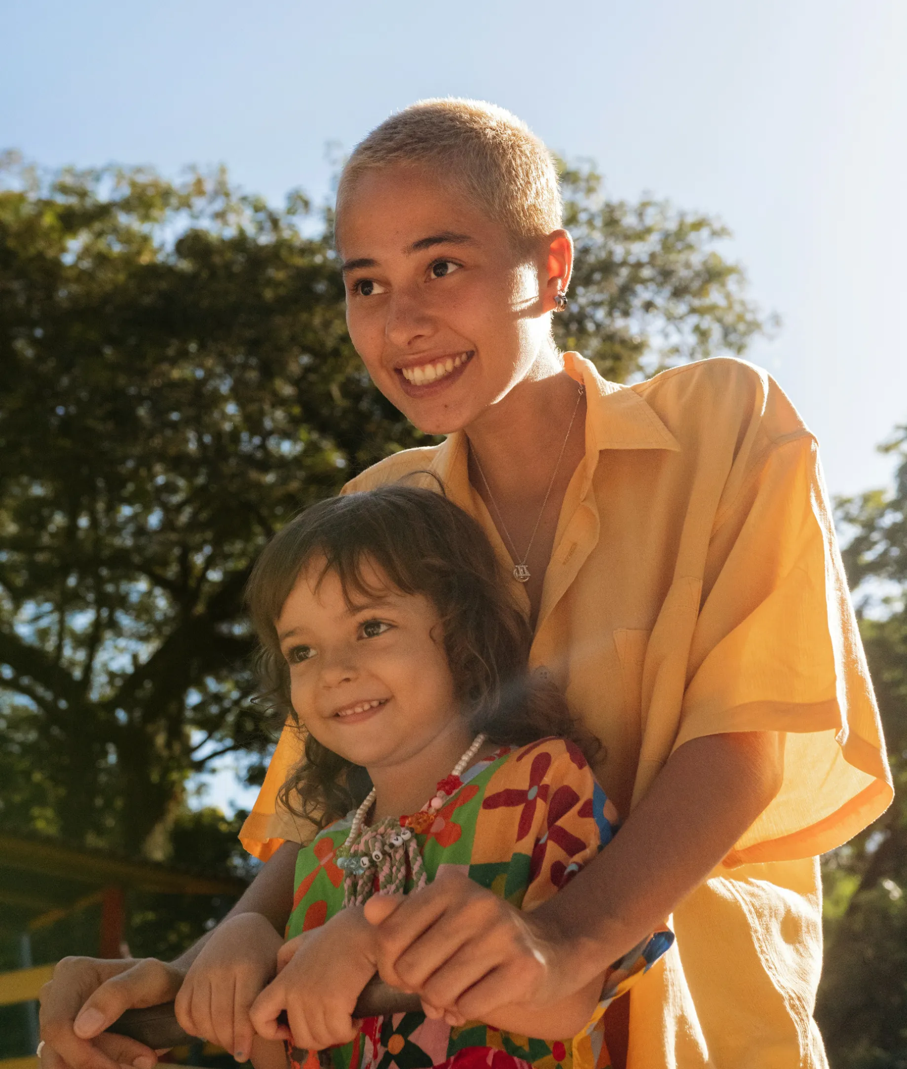 Smiling young person with short blond hair in a yellow shirt embraces a child wearing a colorful dress outdoors with trees in the background.