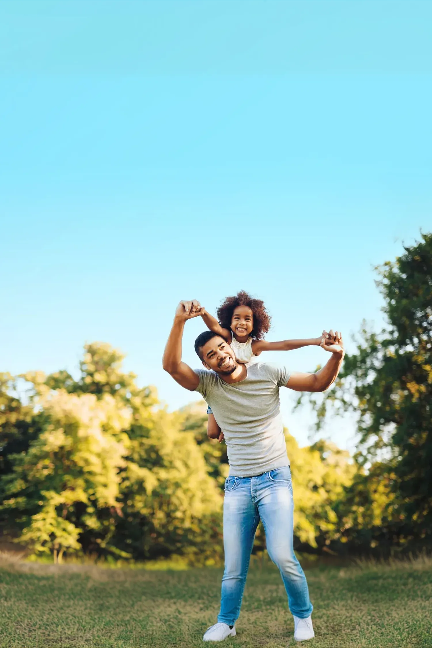 Smiling man carrying a joyful young girl on his back outdoors with trees and blue sky in the background.