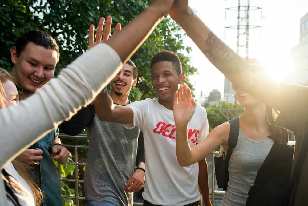 Group of diverse young people outdoors giving high fives and smiling in sunlight.