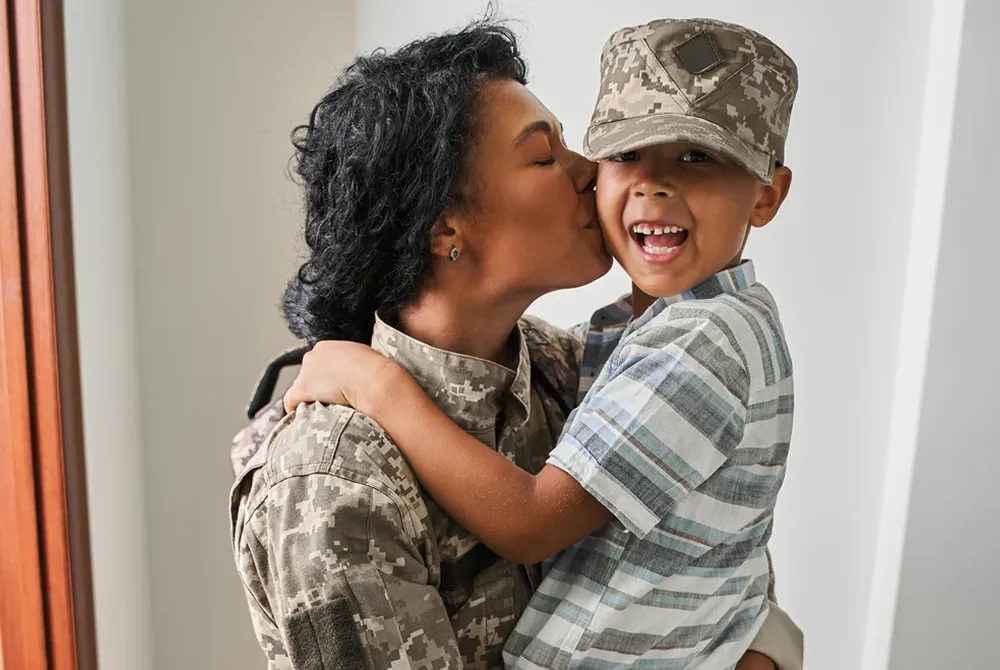 Woman in military uniform kissing and holding a smiling young boy wearing a camouflage cap and striped shirt.