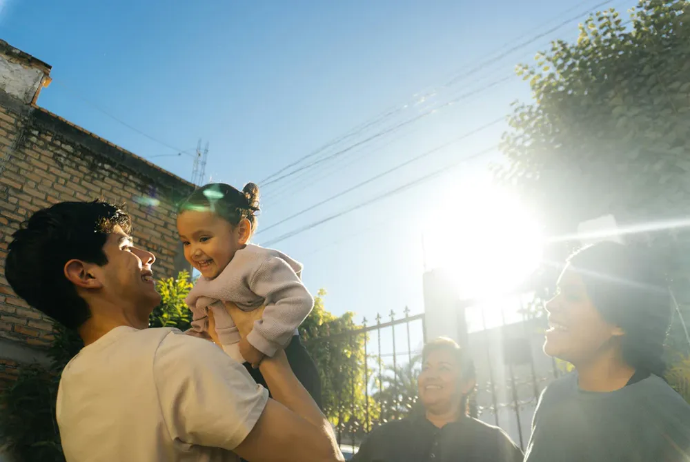 Man lifting and smiling at a happy toddler girl outdoors with two women watching and smiling in bright sunlight.