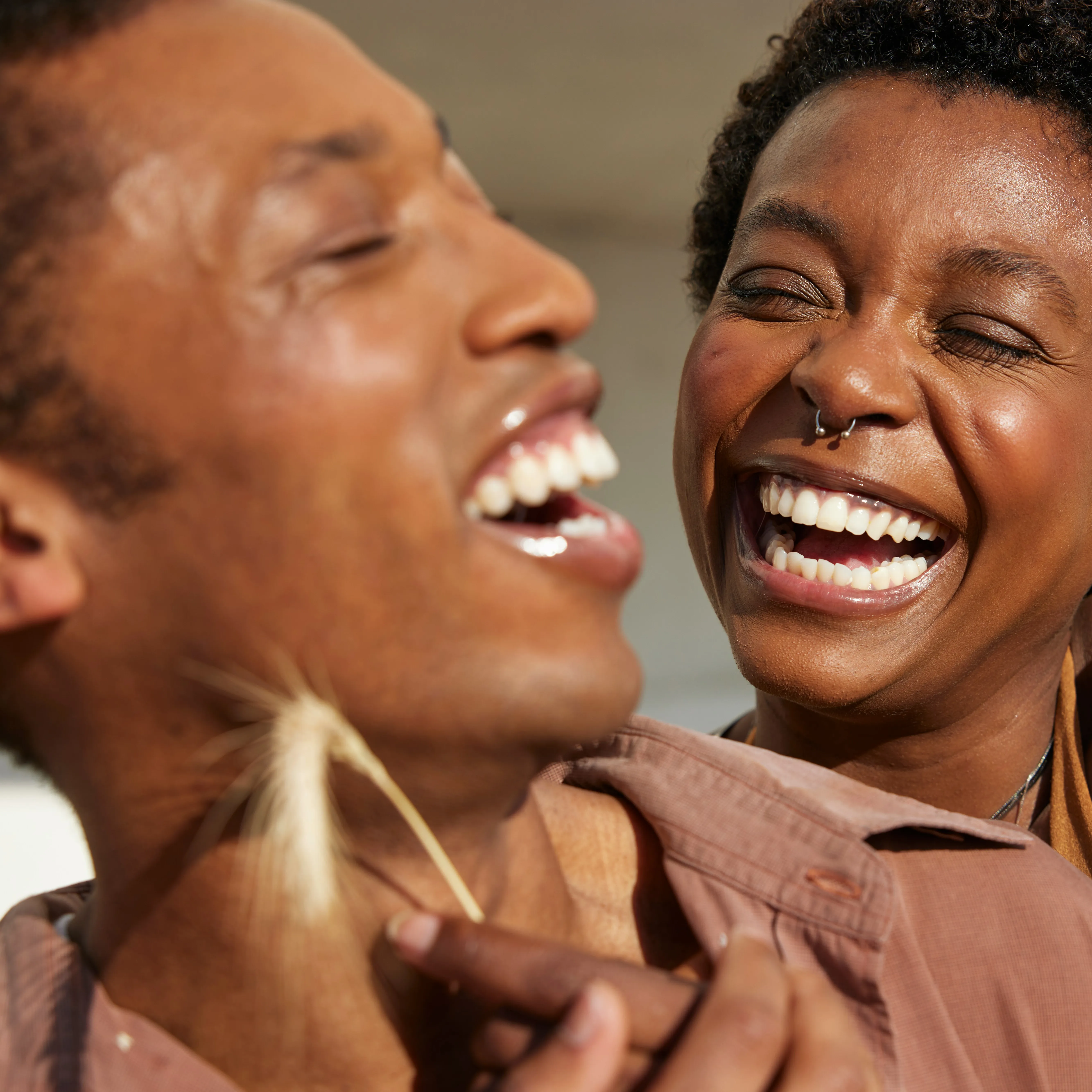 Close-up of two people laughing joyfully, one holding a piece of dried grass near the other's neck.
