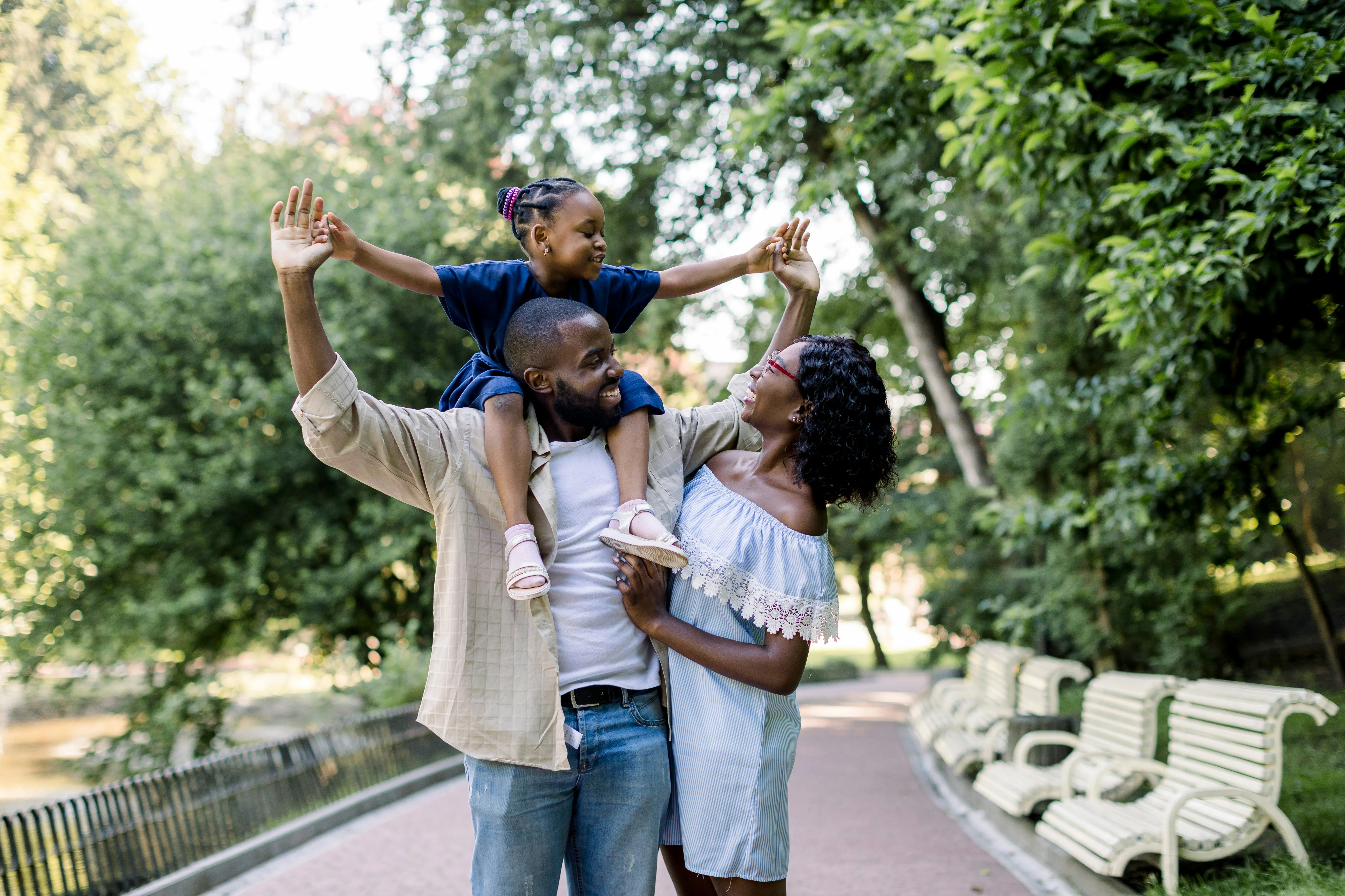 Smiling family with a man carrying a young girl on his shoulders holding hands with a woman on a tree-lined park path.
