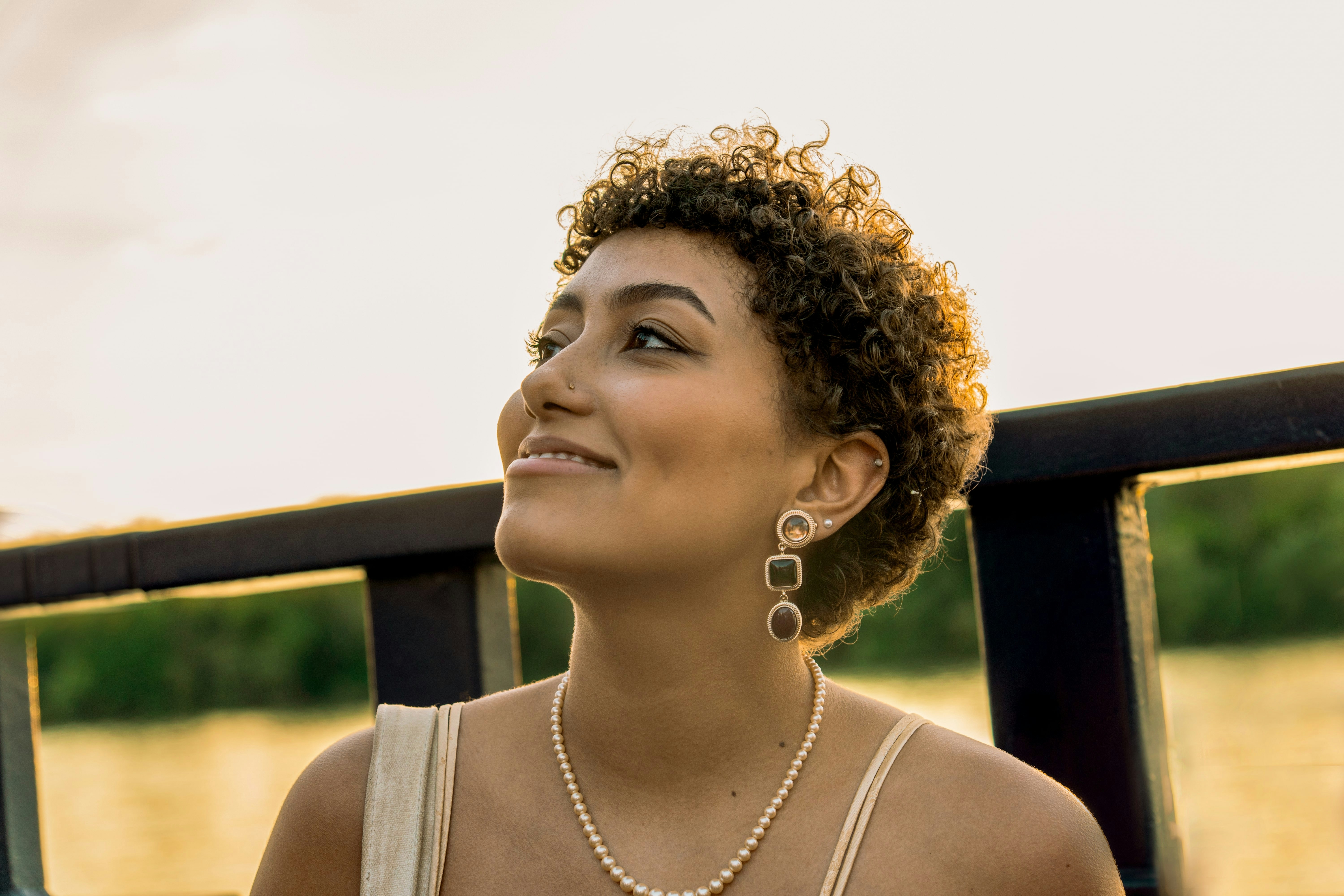 Smiling woman with short curly hair wearing pearl necklace and dangling earrings looking to the side outdoors near water.