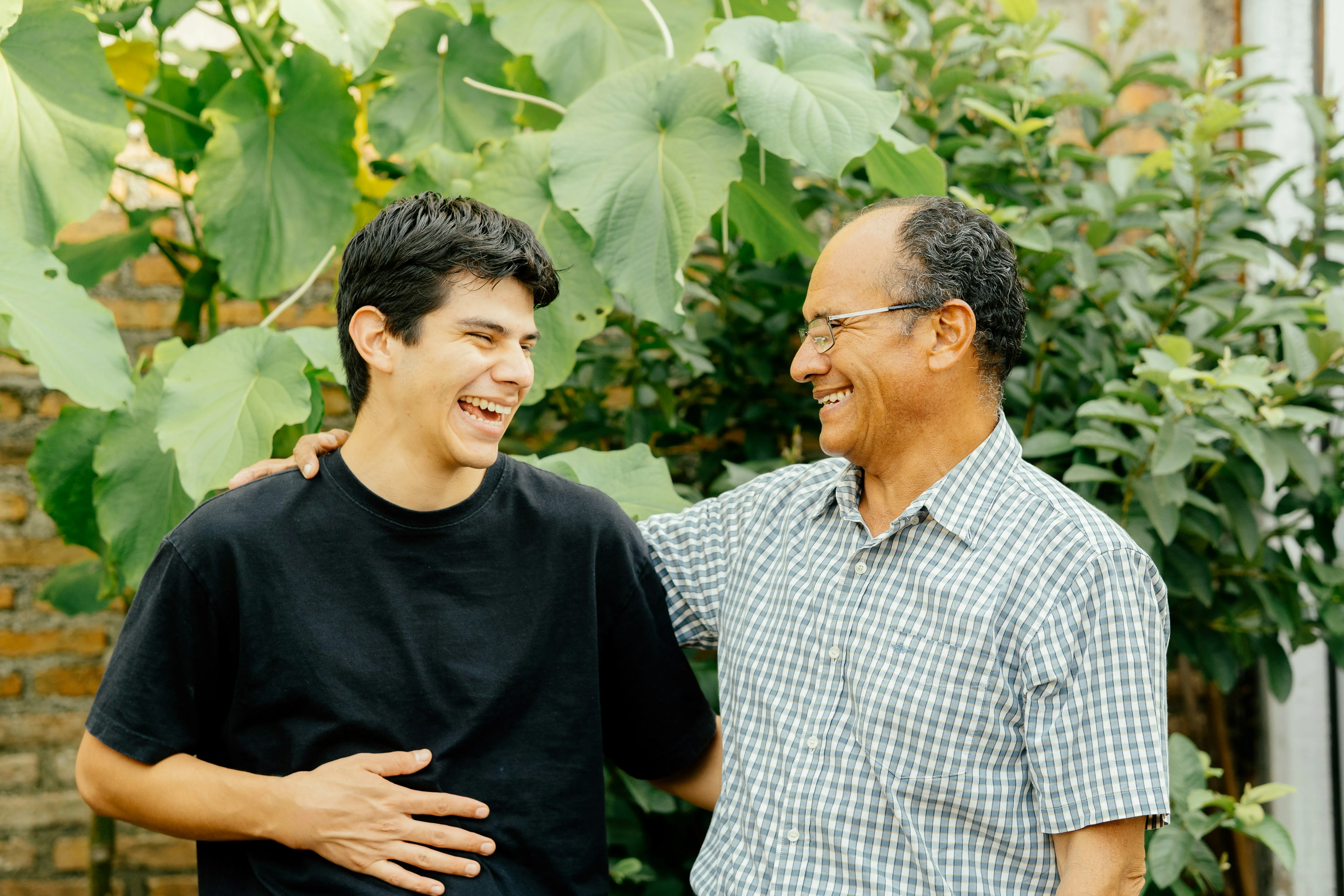 An older man in glasses and a checkered shirt laughing and holding a younger man in a black t-shirt in a garden setting.
