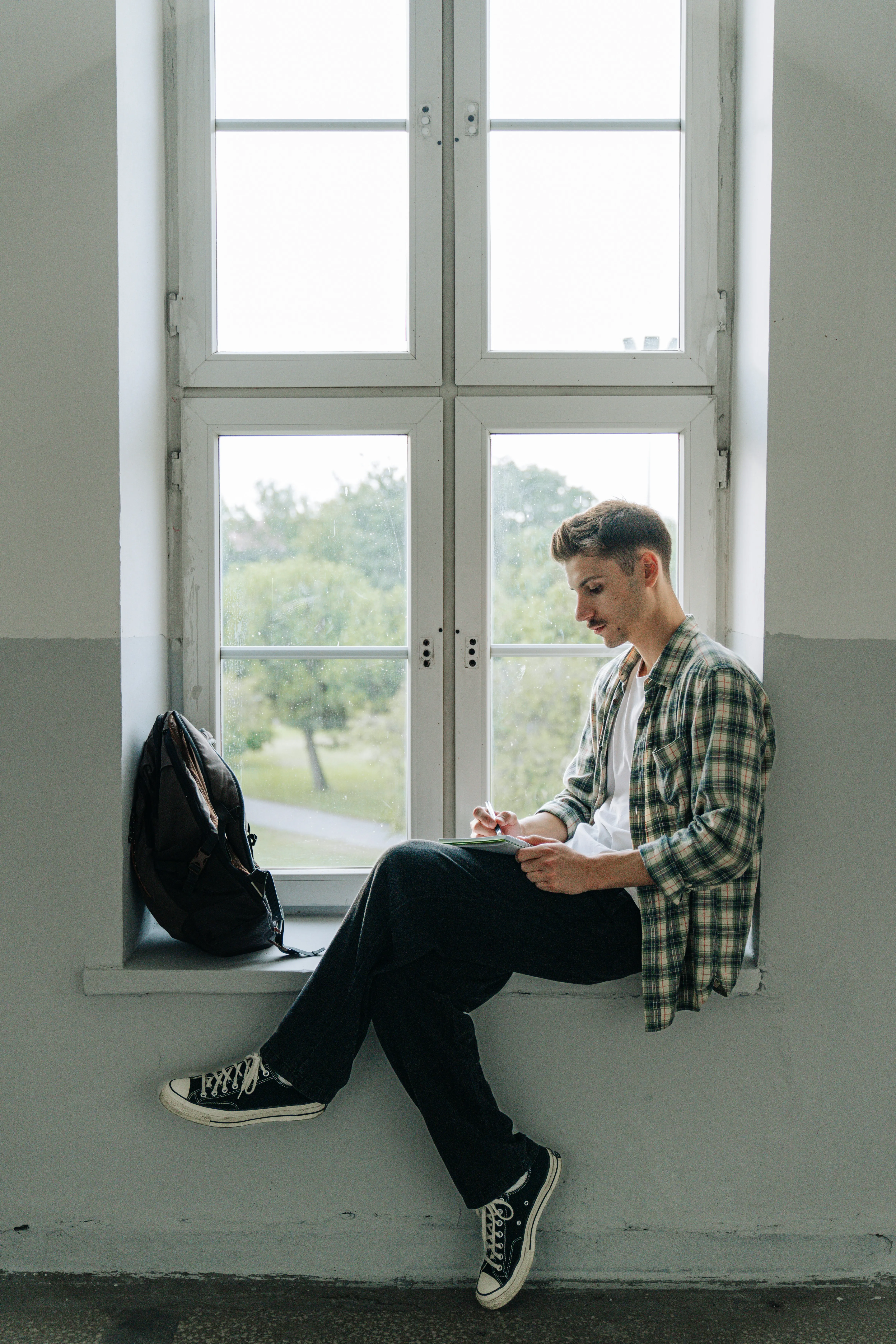 Young man in a plaid shirt sitting on a window ledge, writing in a notebook with a backpack beside him.
