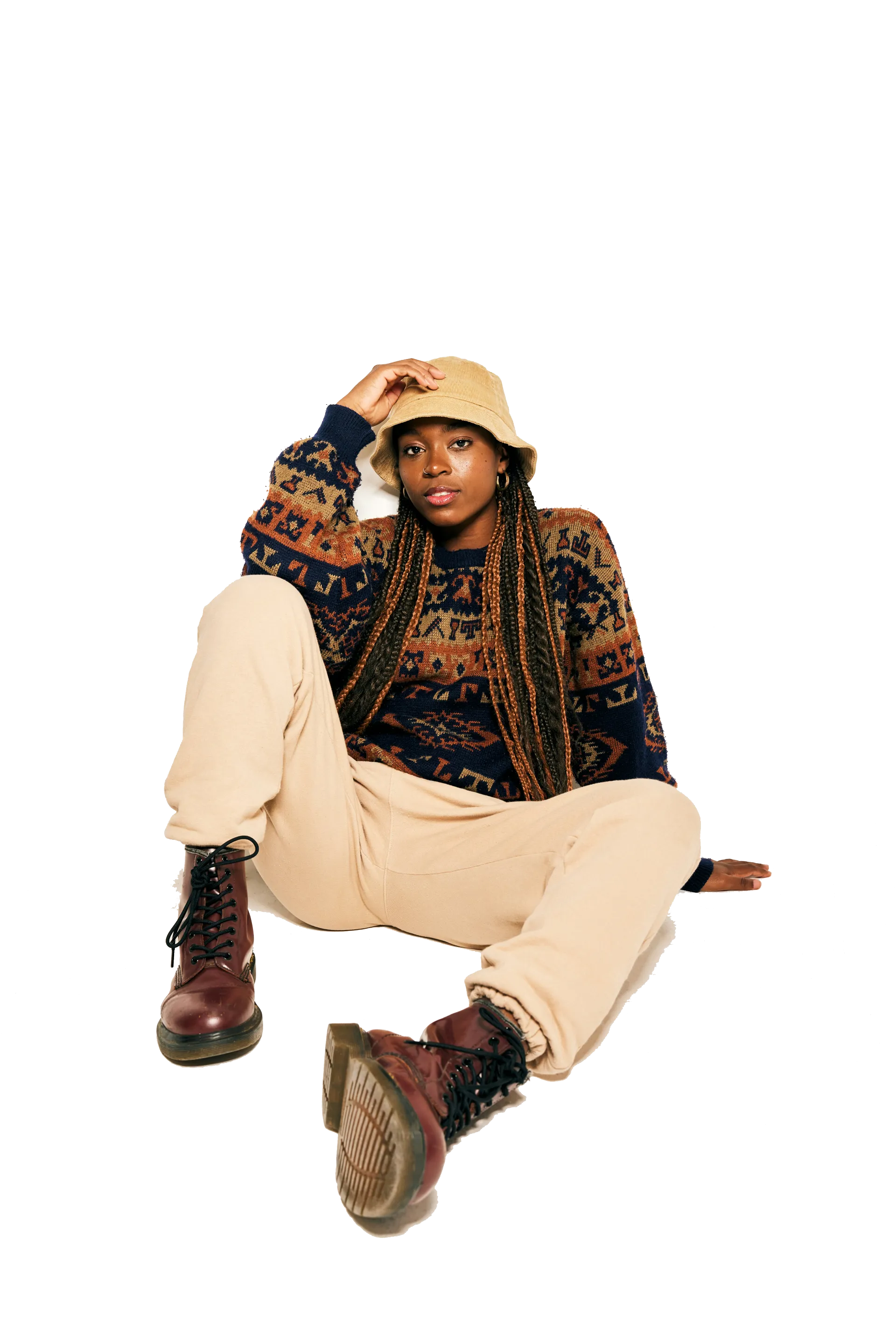 Woman with long braids wearing a patterned sweater, beige pants, bucket hat, and burgundy boots sitting on the floor with one hand touching the hat.