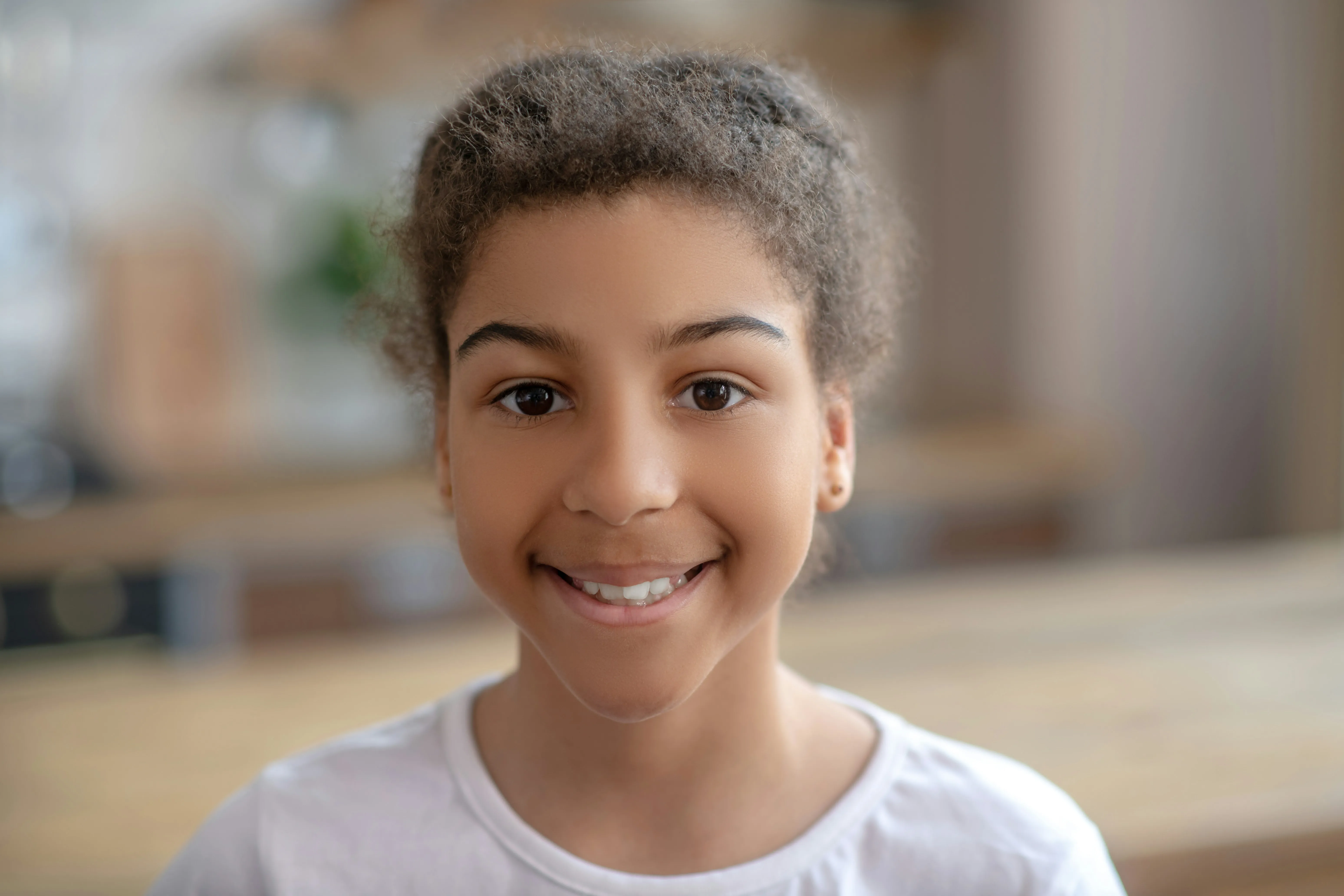 Smiling young girl with curly hair wearing a white shirt in a softly blurred indoor setting.