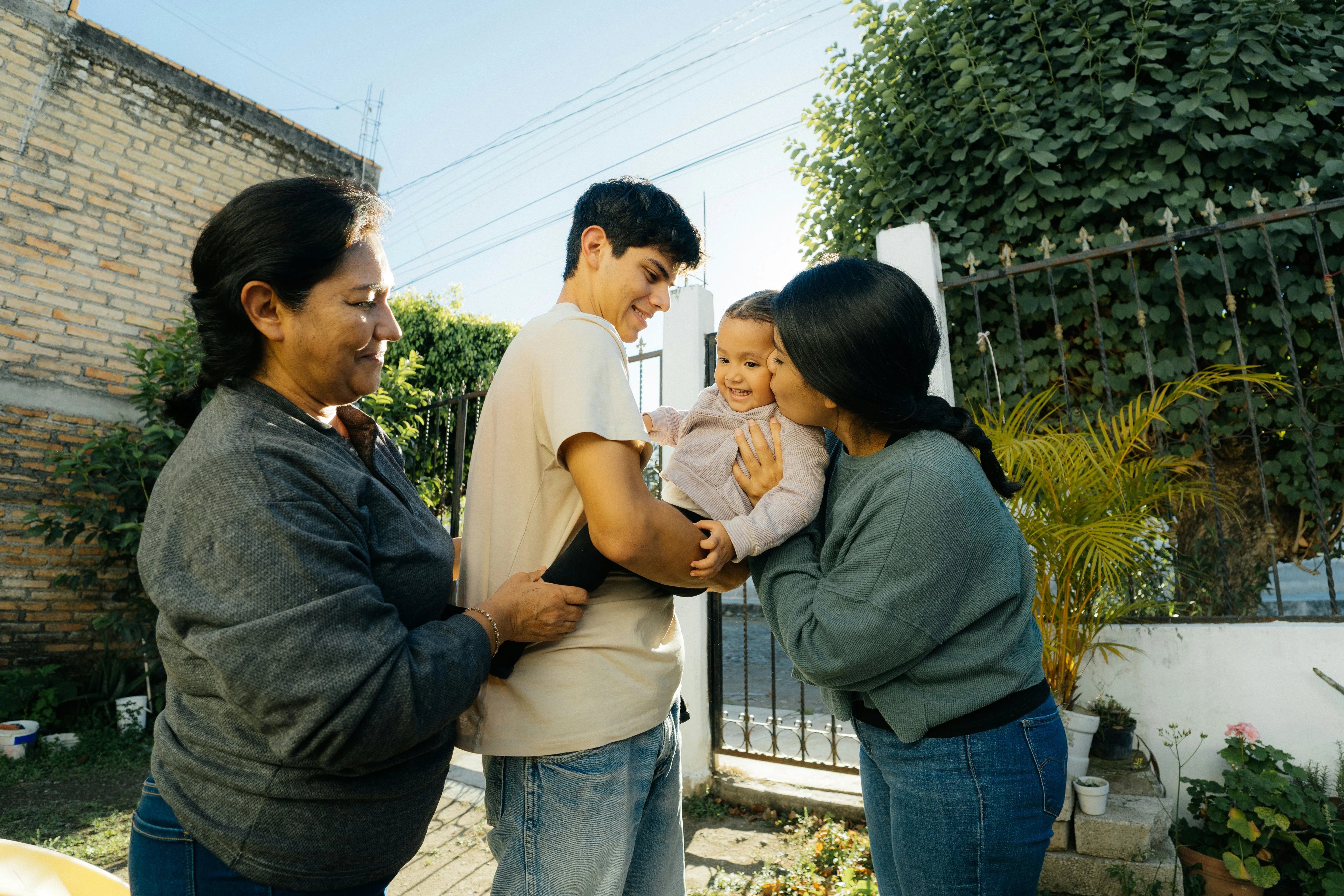 Three adults happily holding and kissing a smiling toddler outdoors near a garden gate.