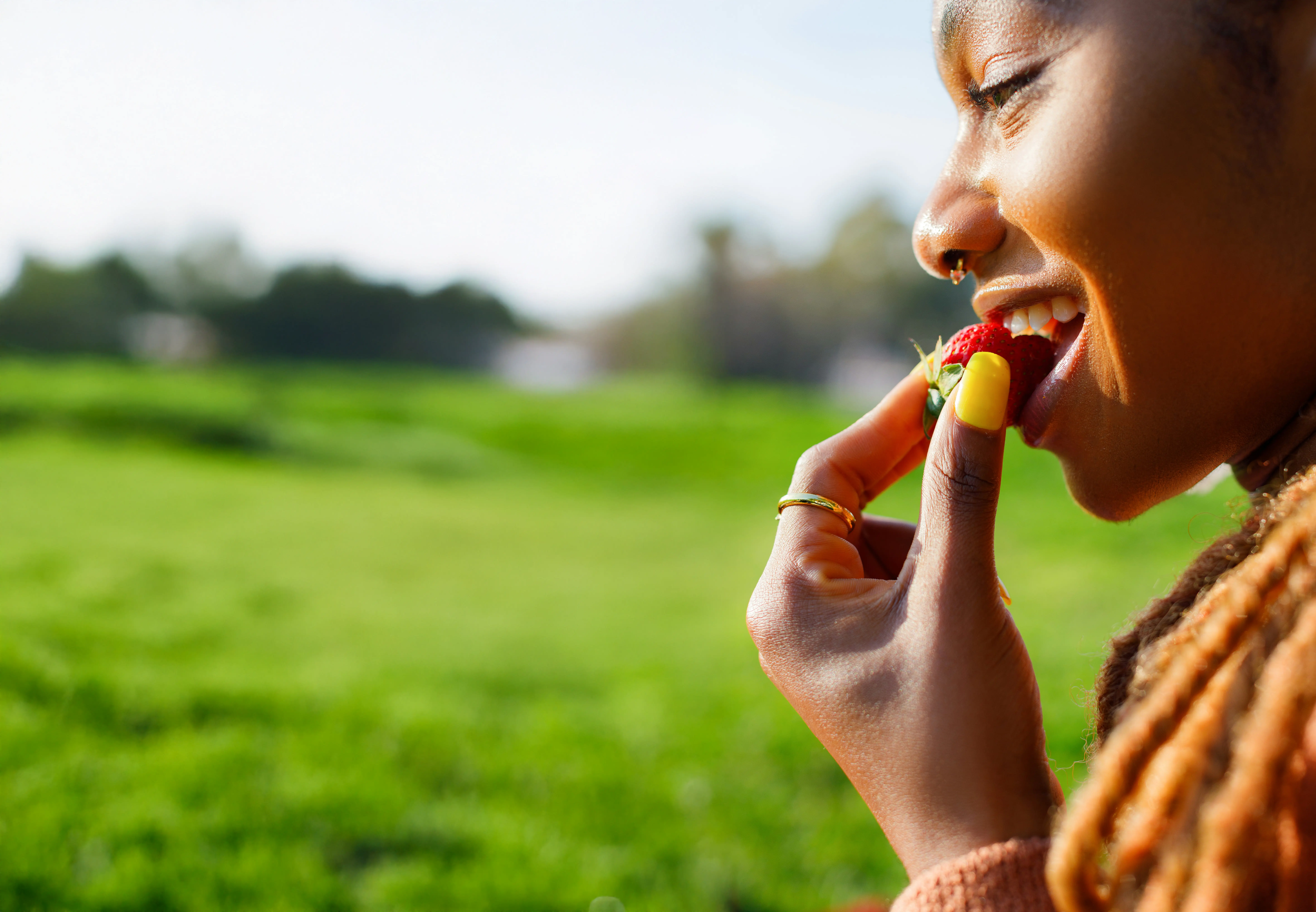 Close-up of a smiling person about to eat a strawberry outdoors with green grass in the background.