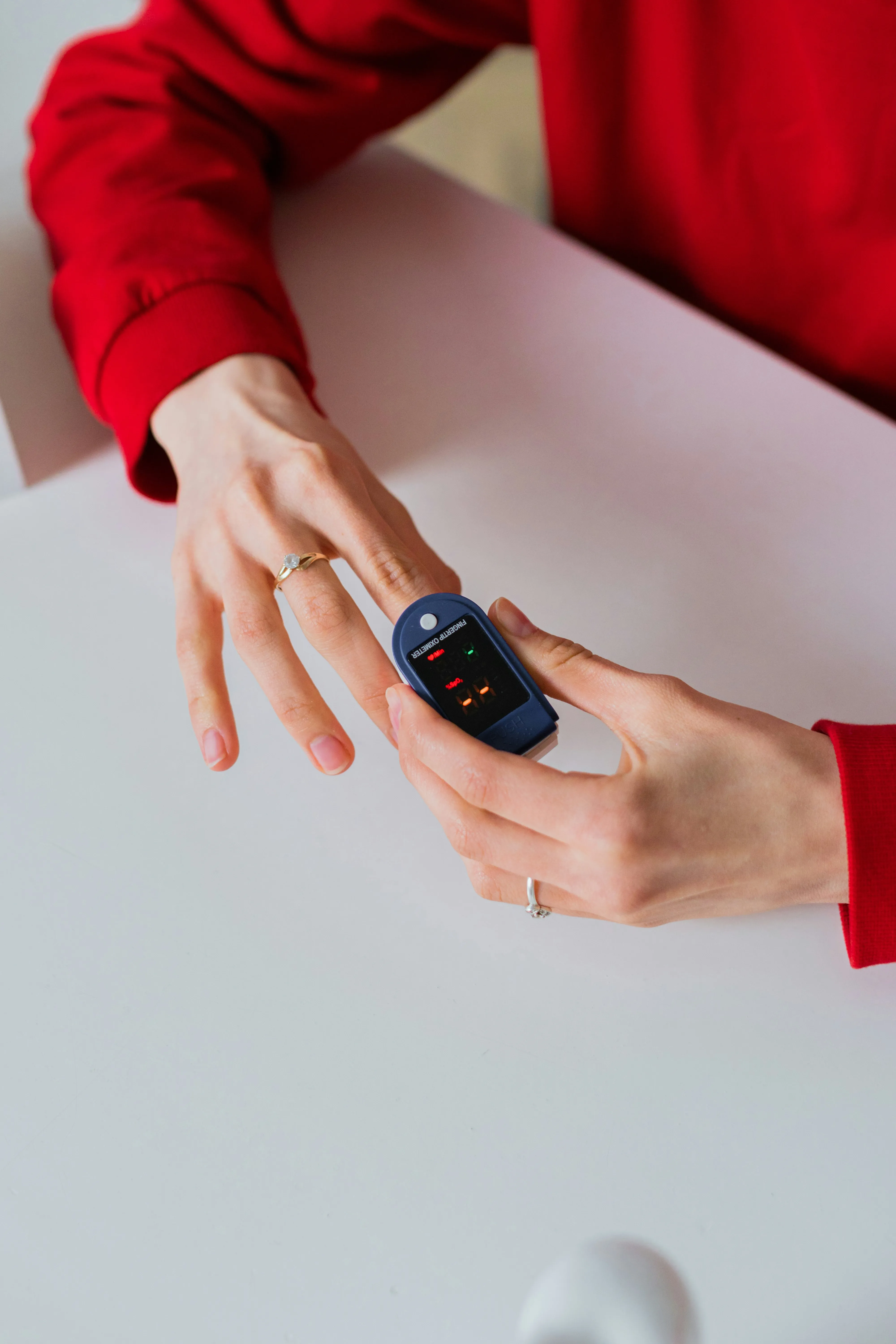 Person wearing red uses a fingertip pulse oximeter to check blood oxygen levels on a finger.