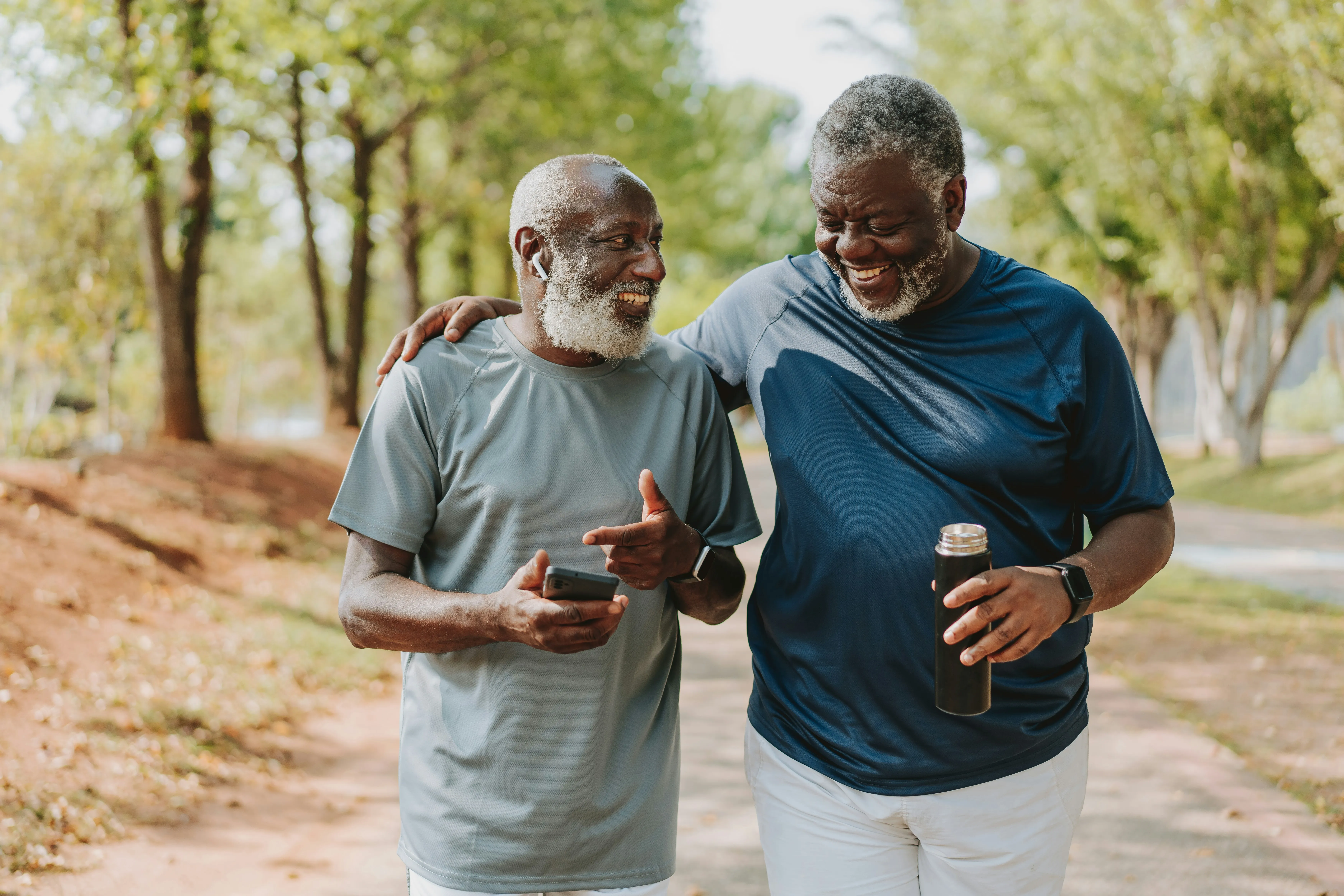 Two older men walking outdoors, smiling, one holding a phone, the other holding a water bottle.