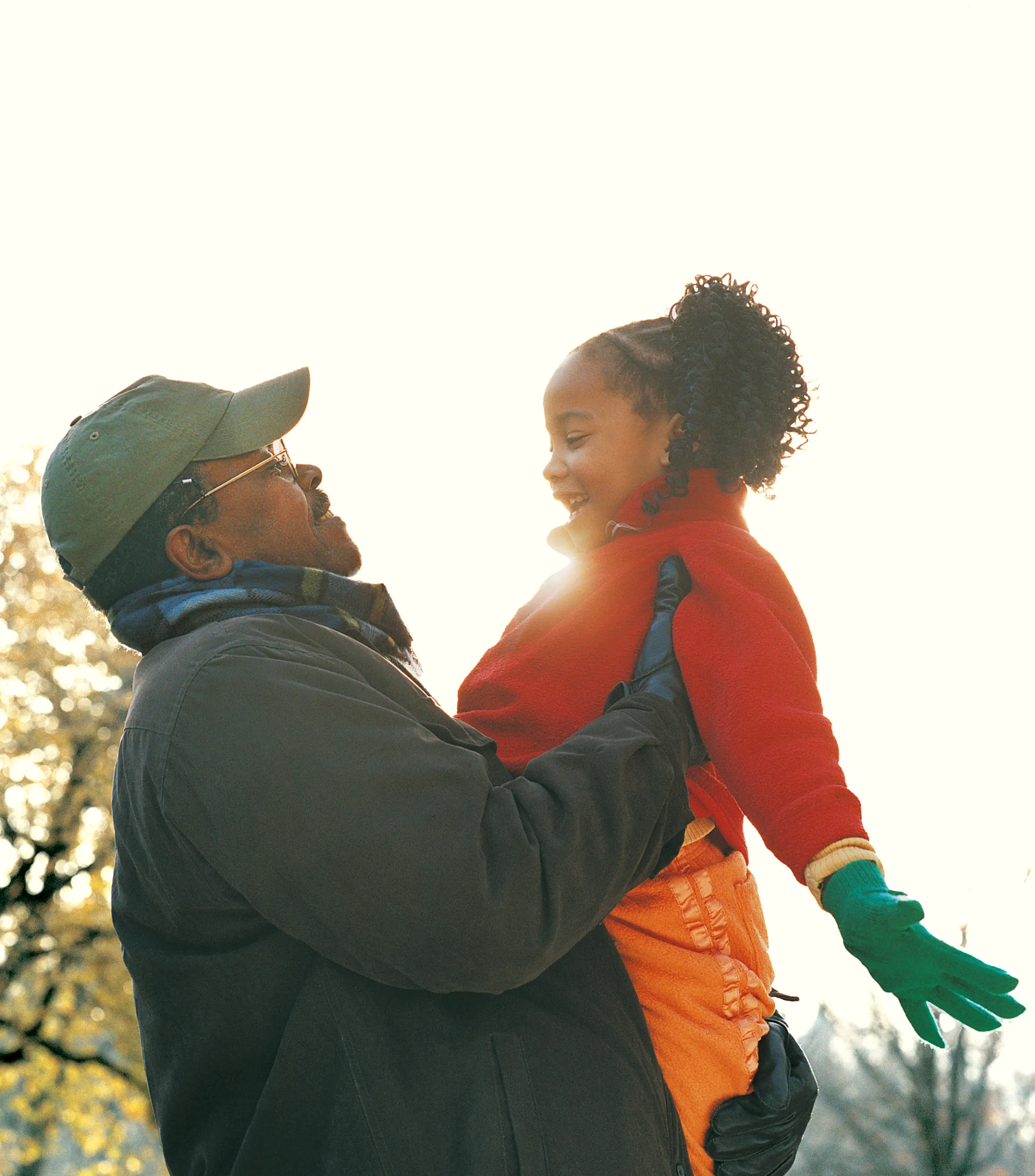 Man in winter clothes lifts a smiling girl wearing green gloves and a red jacket outdoors.