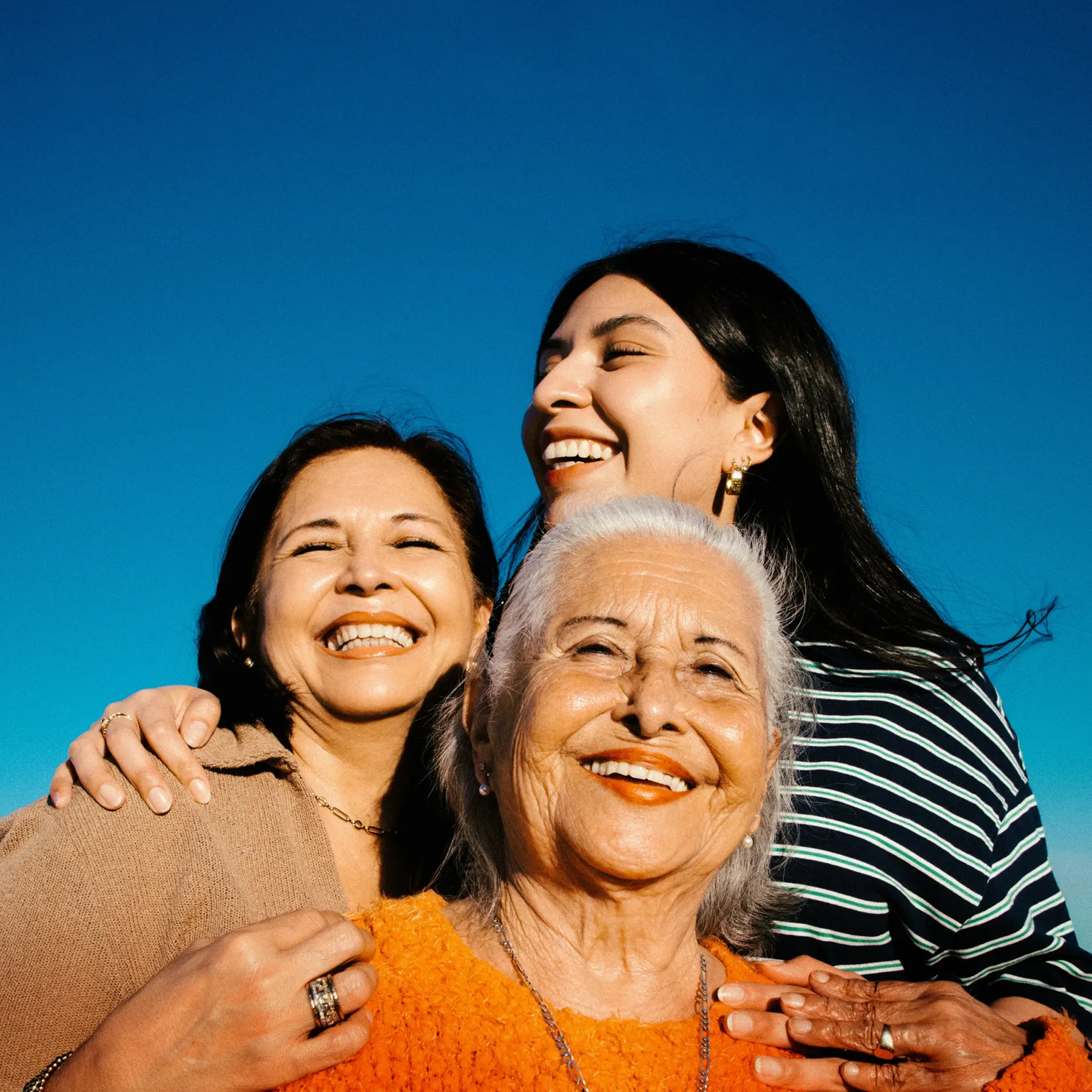 Three happy women of different generations smiling together under a clear blue sky.
