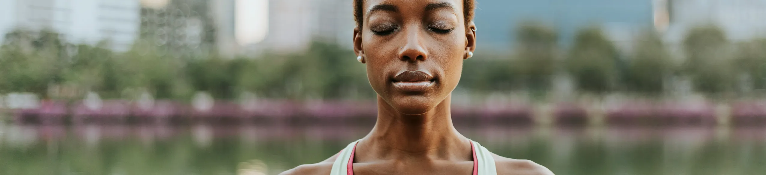 Close-up of a woman with closed eyes meditating outdoors near water and trees.