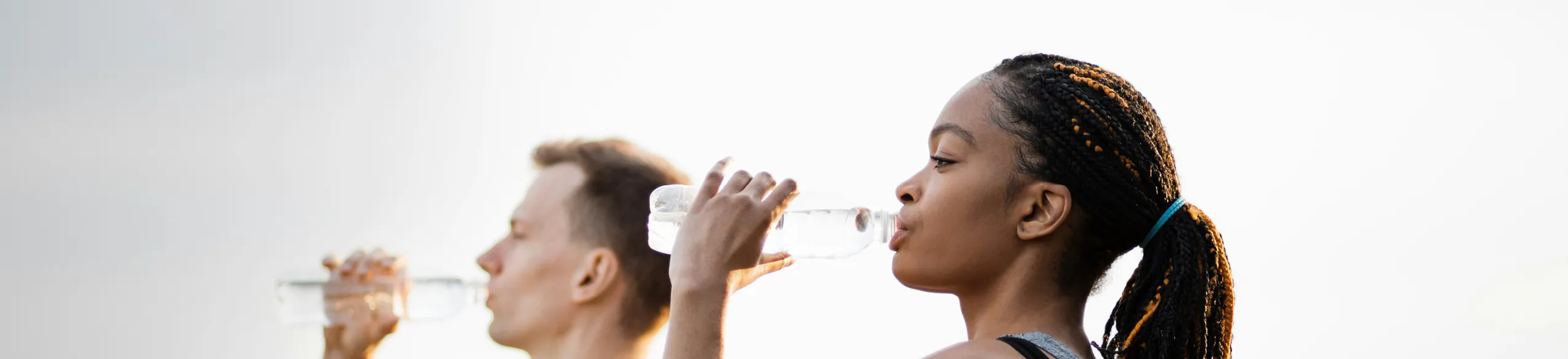 Two people drinking water from plastic bottles outdoors with a bright sky background.