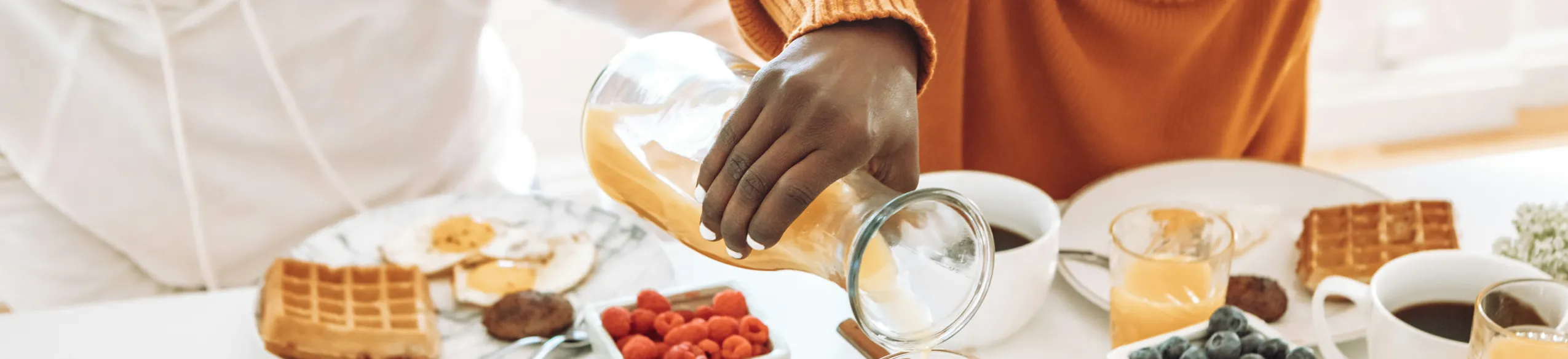 Person in orange sweater pouring juice at a breakfast table with waffles, berries, and coffee.