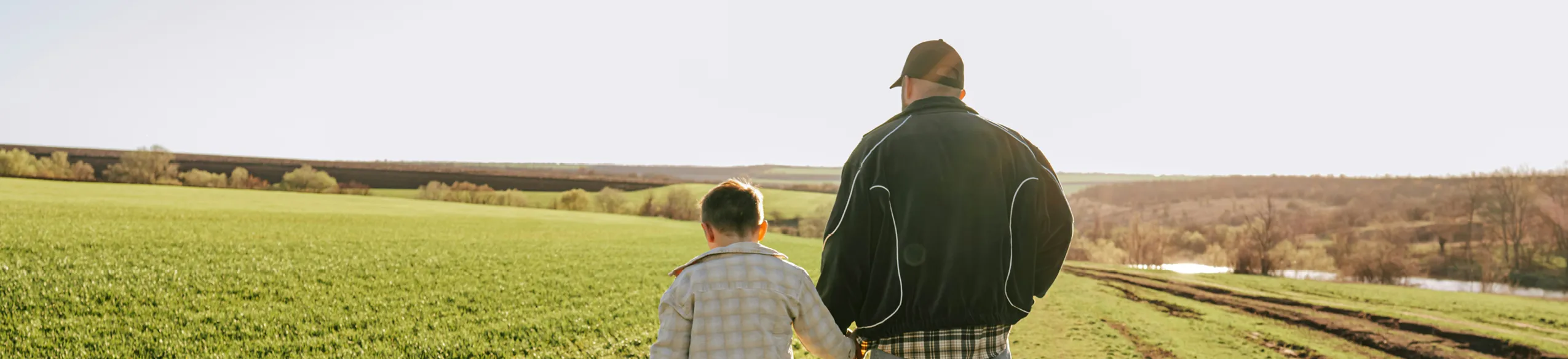 Man and child walking hand in hand through a sunlit grassy field with trees and hills in the distance.