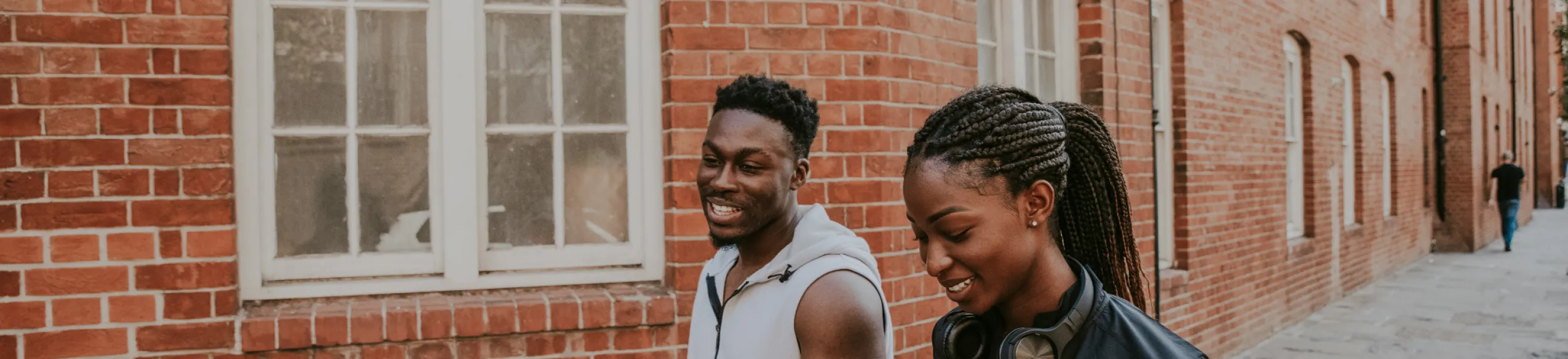Two young people smiling and walking by a brick building with multiple white windows.