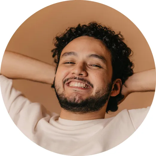 Smiling man with curly hair and beard reclining with hands behind head against a beige background.
