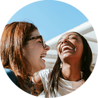 Two women smiling and laughing outdoors against a clear blue sky background.