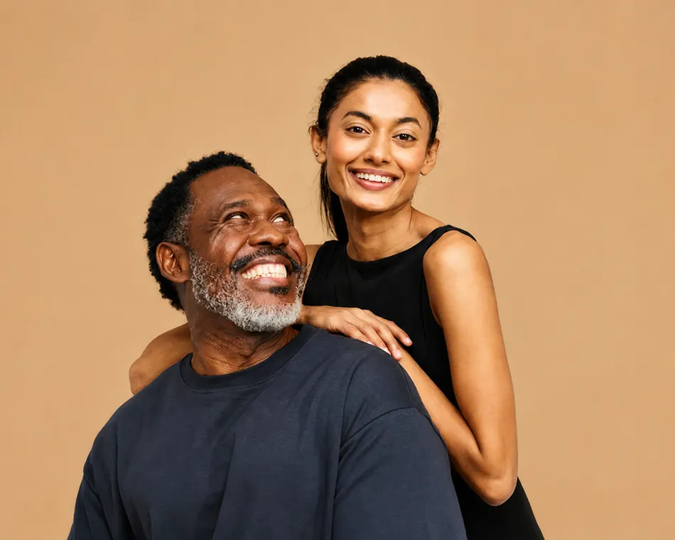 Smiling man with gray beard in dark shirt looks up at smiling woman in black dress behind him.