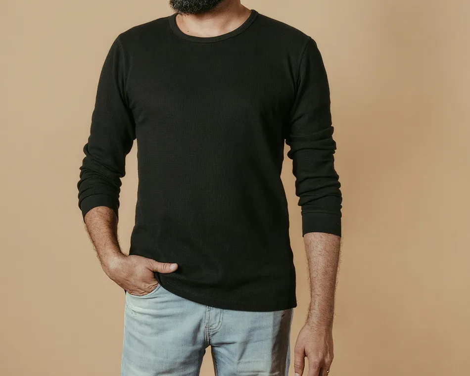 Man with beard wearing a black long-sleeve shirt and light blue jeans standing against beige background.