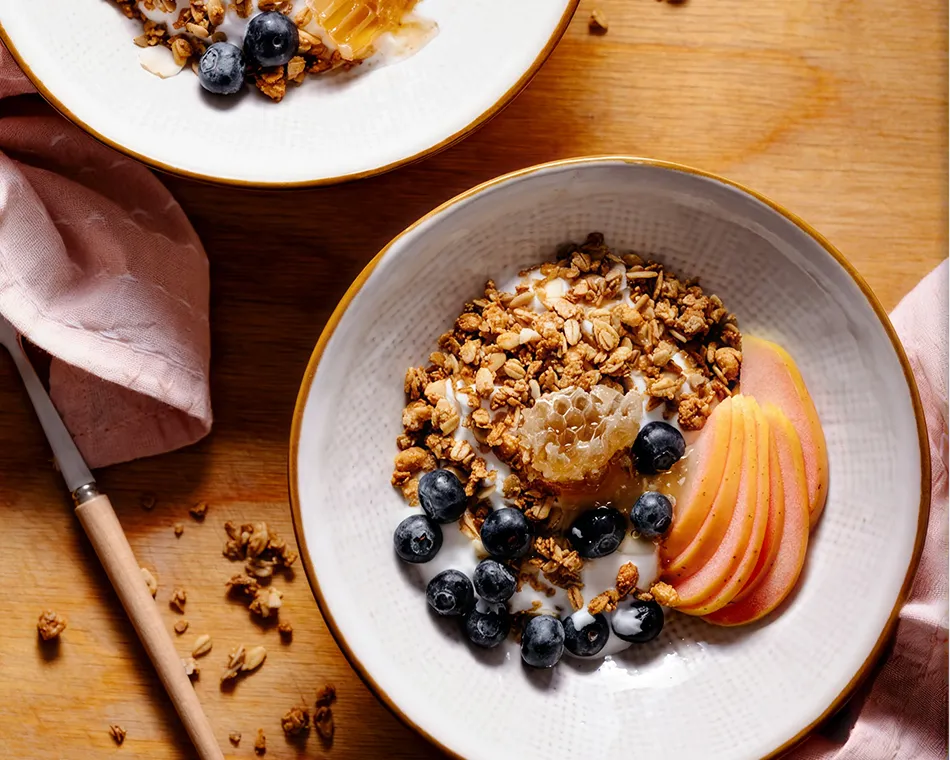 Two bowls of yogurt with granola, blueberries, apple slices, and honeycomb on a wooden surface.