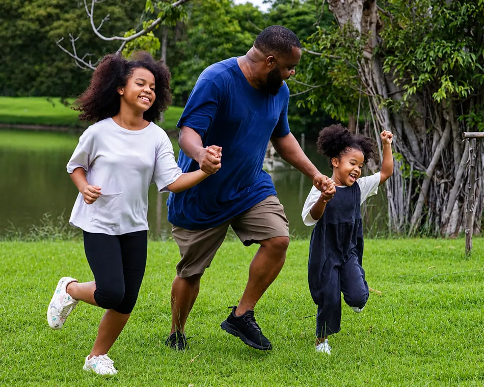 Man holding hands with two young girls as they run happily on grass near trees and water.