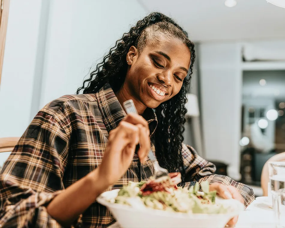Smiling woman in plaid shirt eating salad indoors at a dining table.
