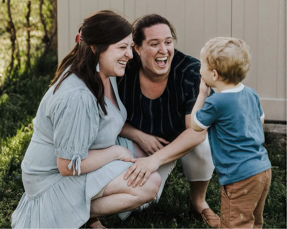 Two women smiling and kneeling while engaging with a young boy outdoors near a building wall.