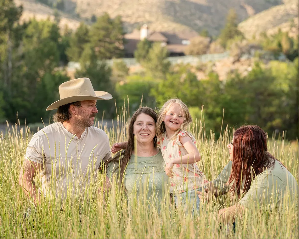 Family of four smiling in a grassy field with trees and mountains in the background.