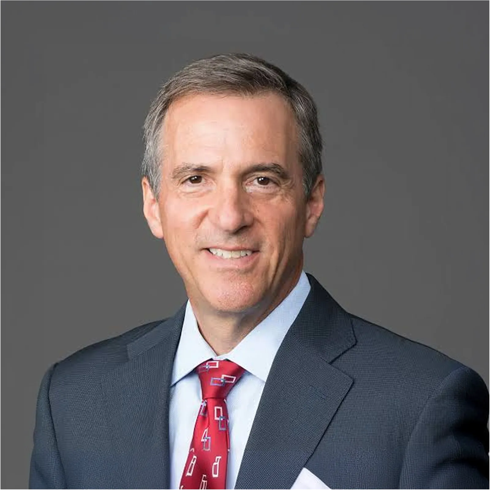 Middle-aged man in a dark suit, light blue shirt, and red patterned tie smiling against a gray background.