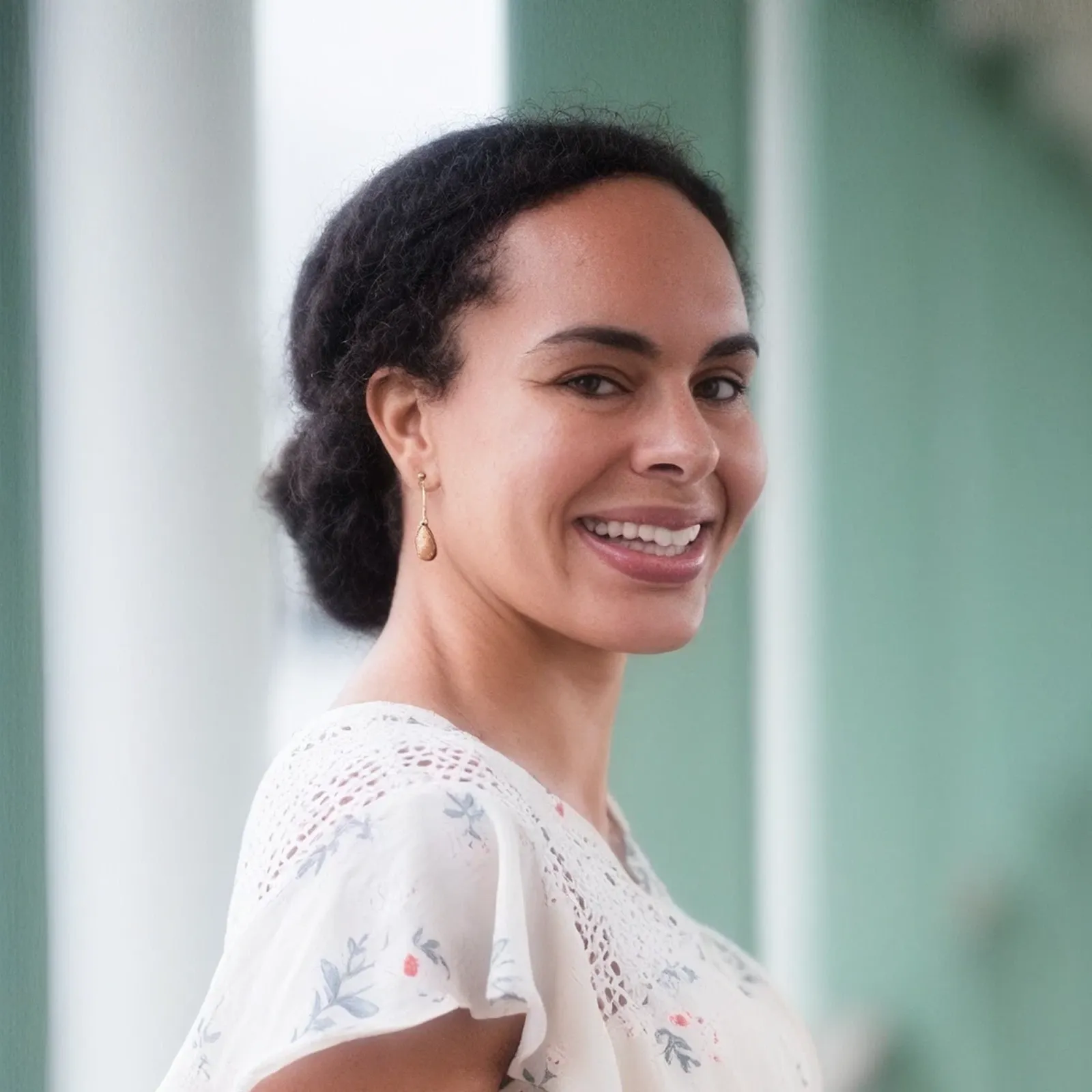 Woman with dark curly hair wearing a white floral blouse and gold drop earrings smiling over her shoulder.