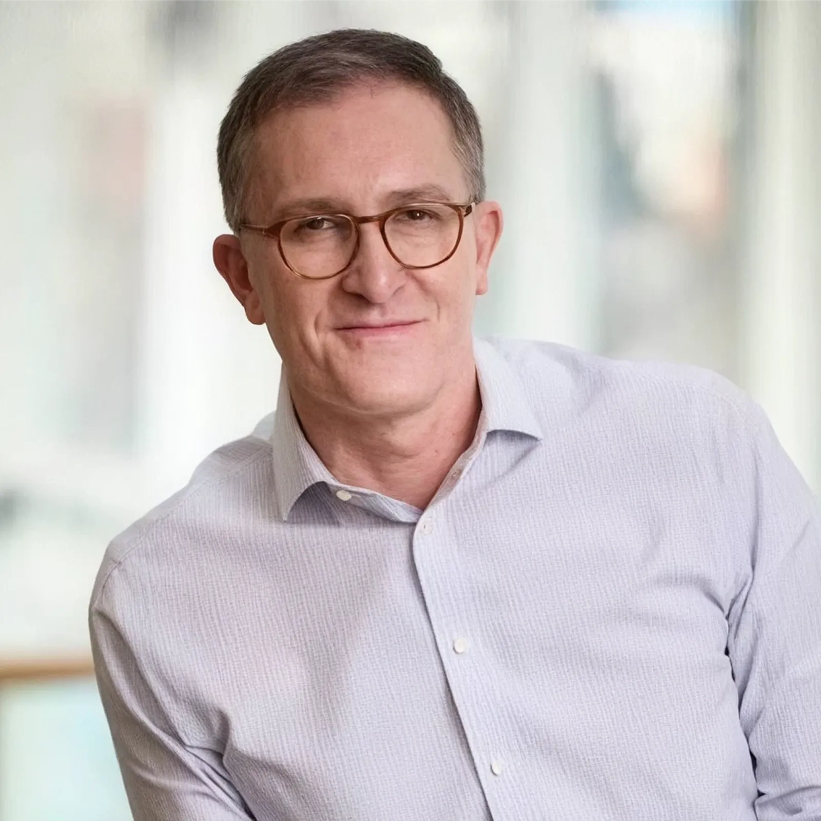 Man wearing glasses and a light gray shirt smiling in a softly lit indoor setting.