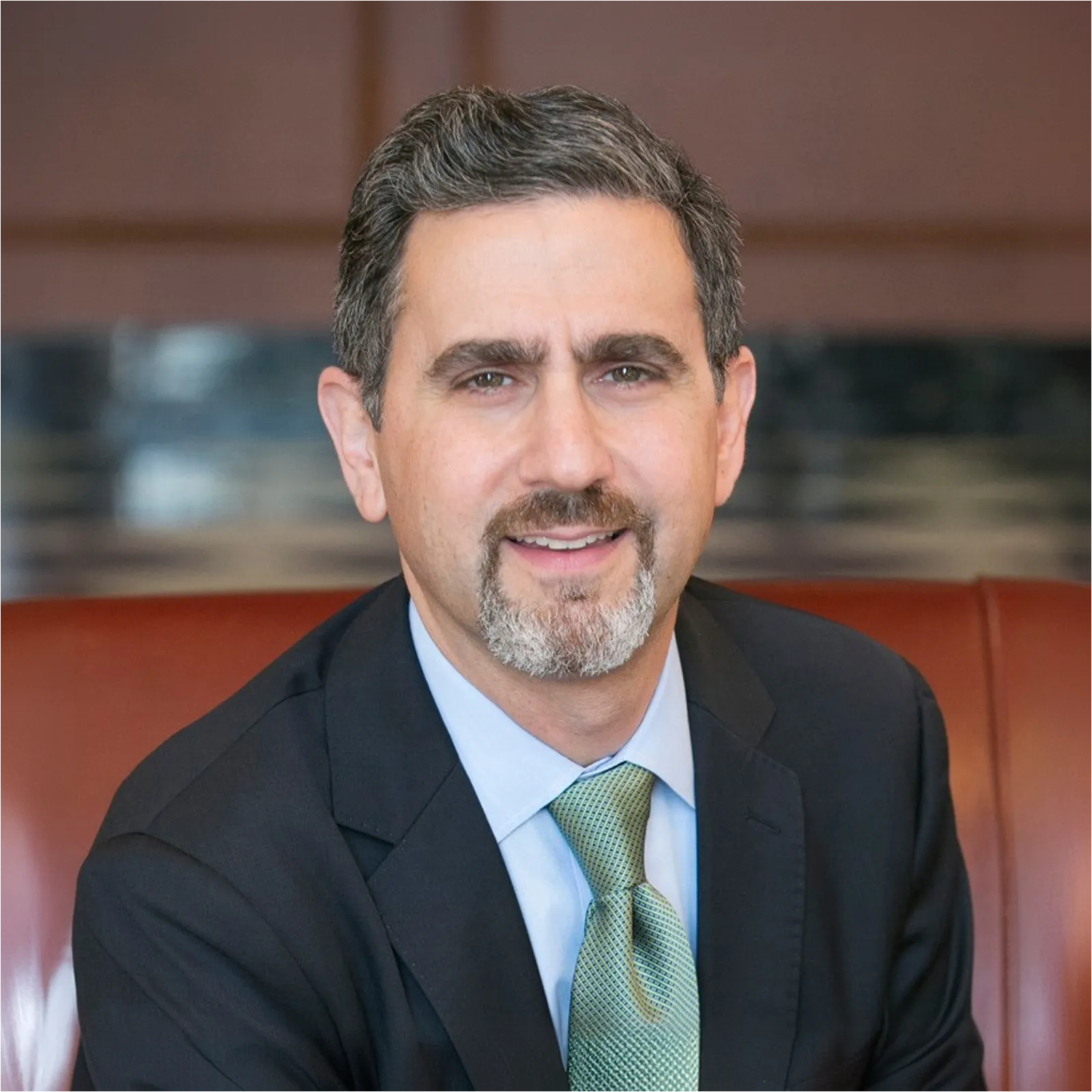 Man with salt-and-pepper hair and beard wearing a dark suit and green tie, smiling seated indoors.