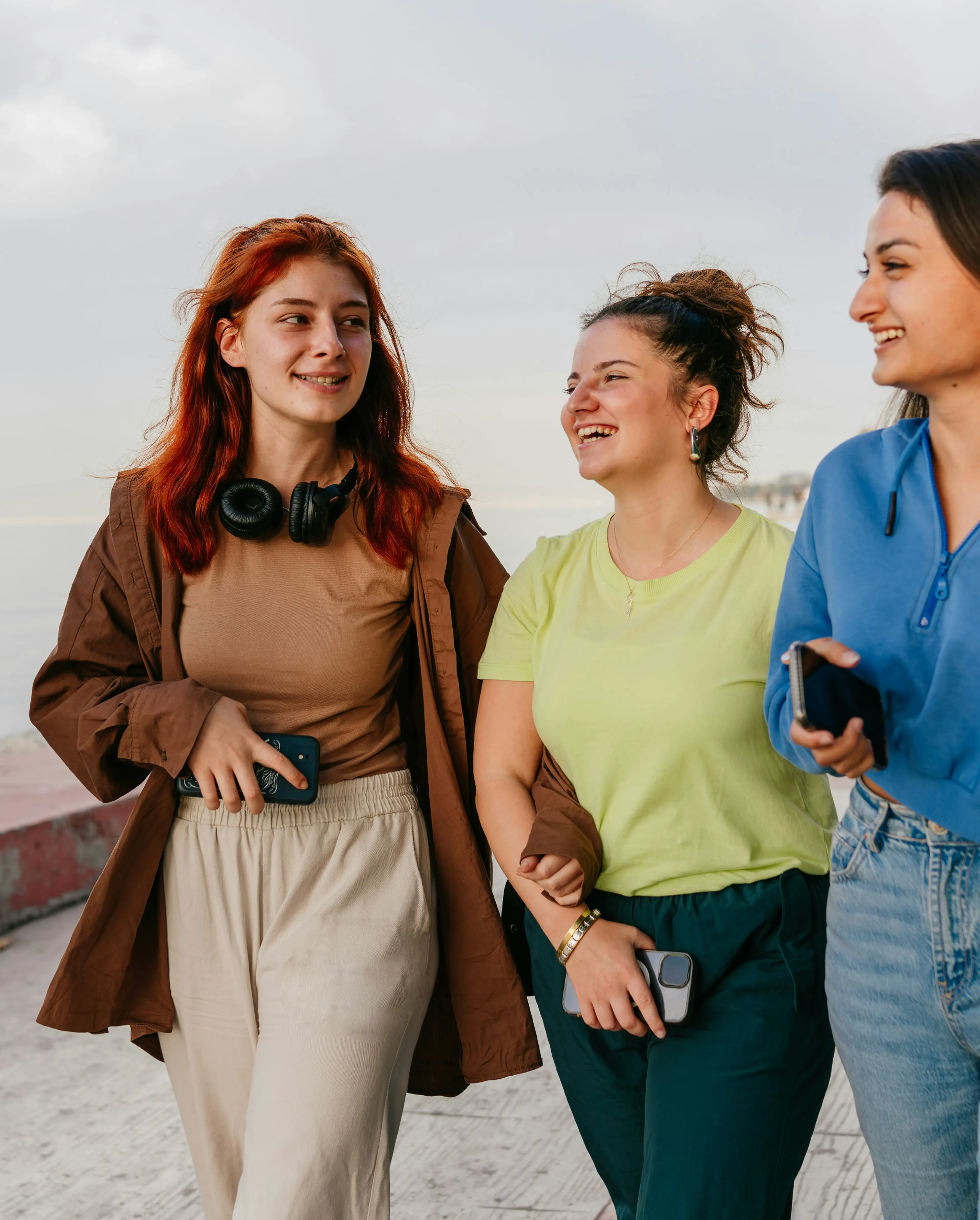 Three young women walking outside near a body of water, smiling and engaged in conversation; one with red hair wearing headphones around her neck, another in a light green shirt holding a phone, and the third in a blue top also holding a phone.