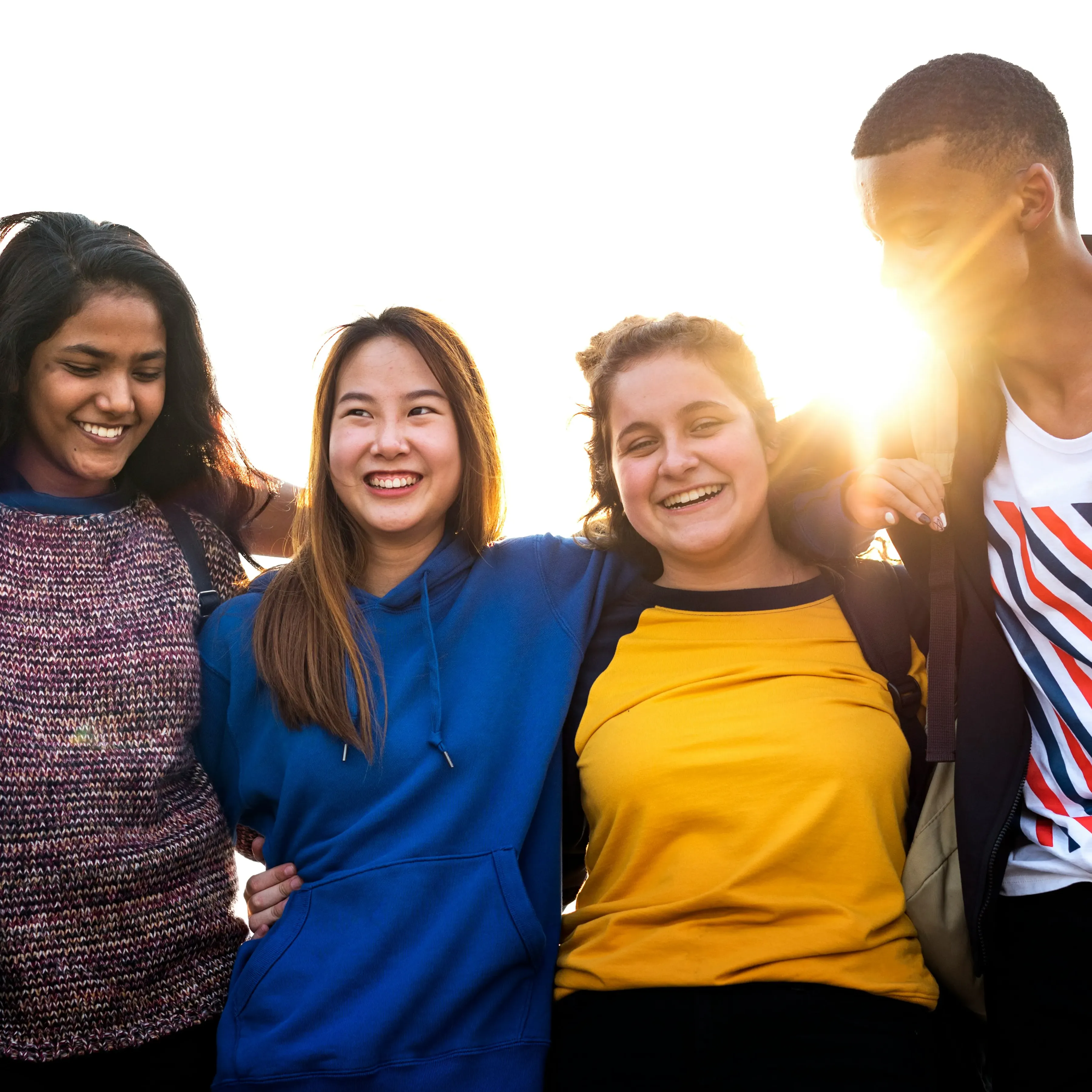 Four diverse young adults standing closely with arms around each other, smiling and laughing with bright sunlight behind them.