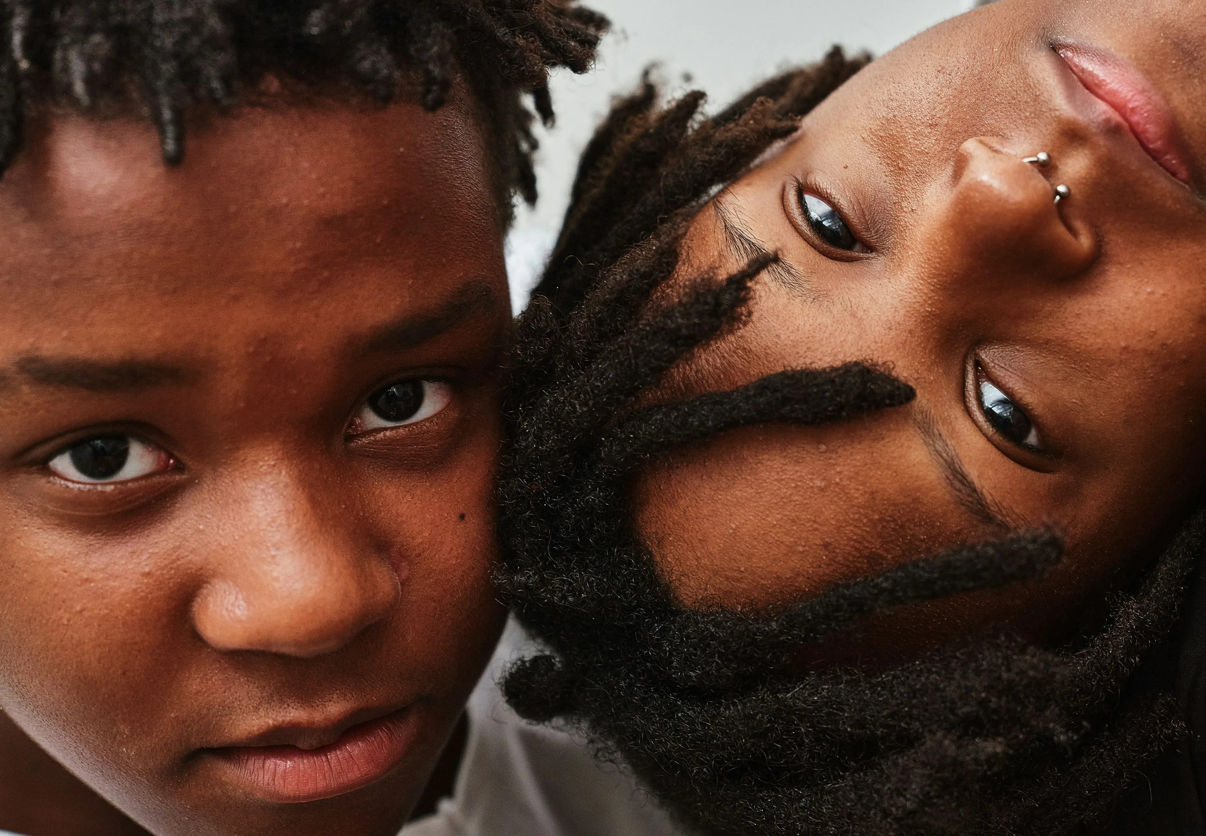 Close-up of two young Black individuals' faces pressed together, one looking directly at the camera with a neutral expression, the other lying sideways with a nose ring and relaxed eyes.