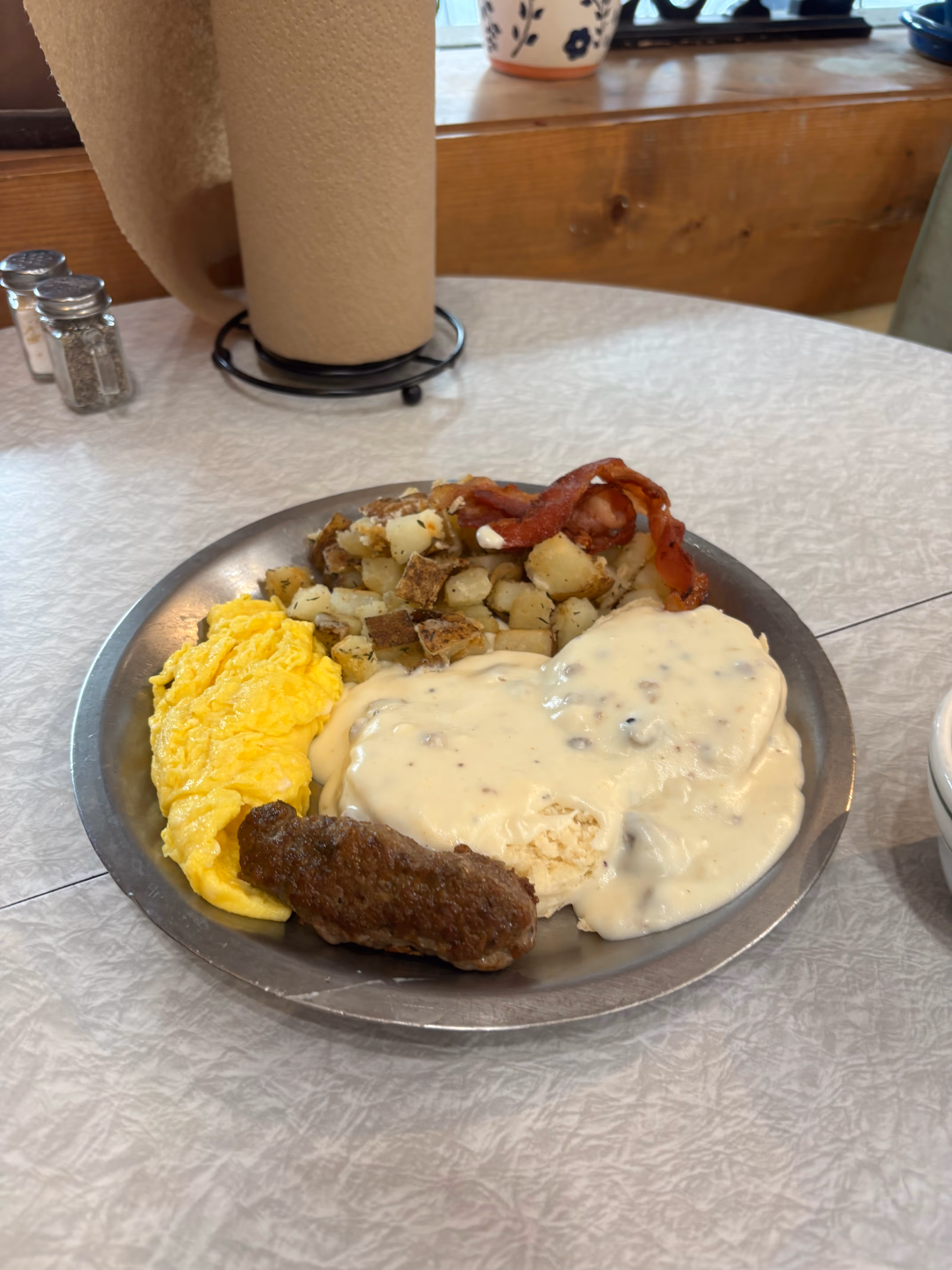Breakfast plate with scrambled eggs, sausage link, bacon strips, diced fried potatoes, and biscuits covered in white gravy.