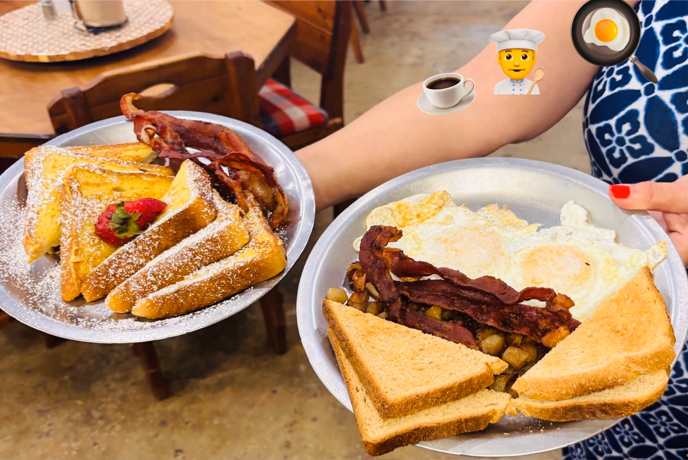 Person holding two plates of breakfast food: one with French toast dusted with powdered sugar topped with a strawberry and bacon strips, the other with two sunny-side-up eggs, two strips of bacon, home fries, and two triangular pieces of toasted bread.