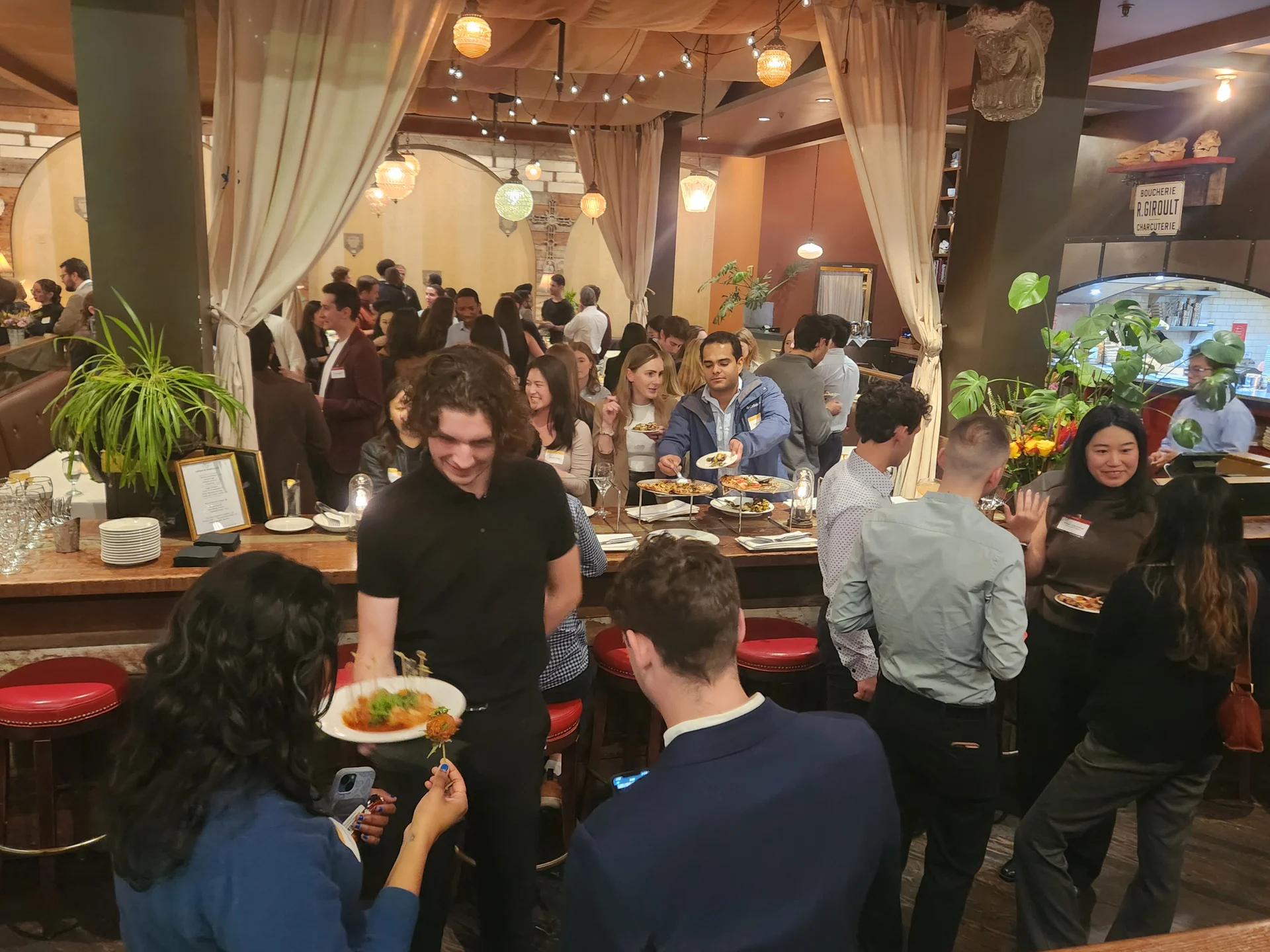 A group of people gathered around a table in a restaurant, enjoying their meals. The setting includes tableware and food, with individuals engaged in conversation and eating. The scene is indoors, featuring walls decorated for the dining experience.