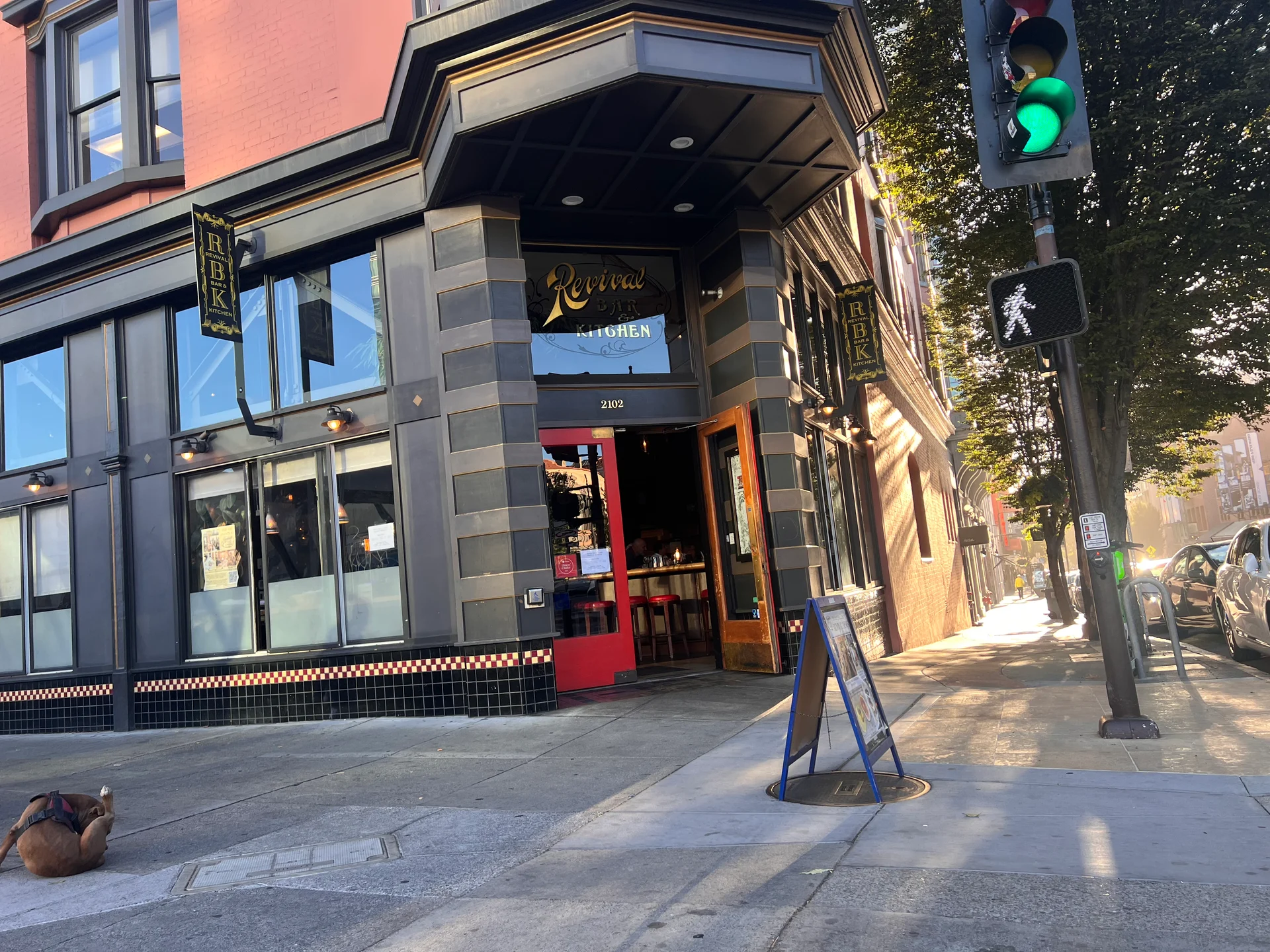 A corner of a building featuring a green traffic light, surrounded by trees and city infrastructure, including windows and a sidewalk.